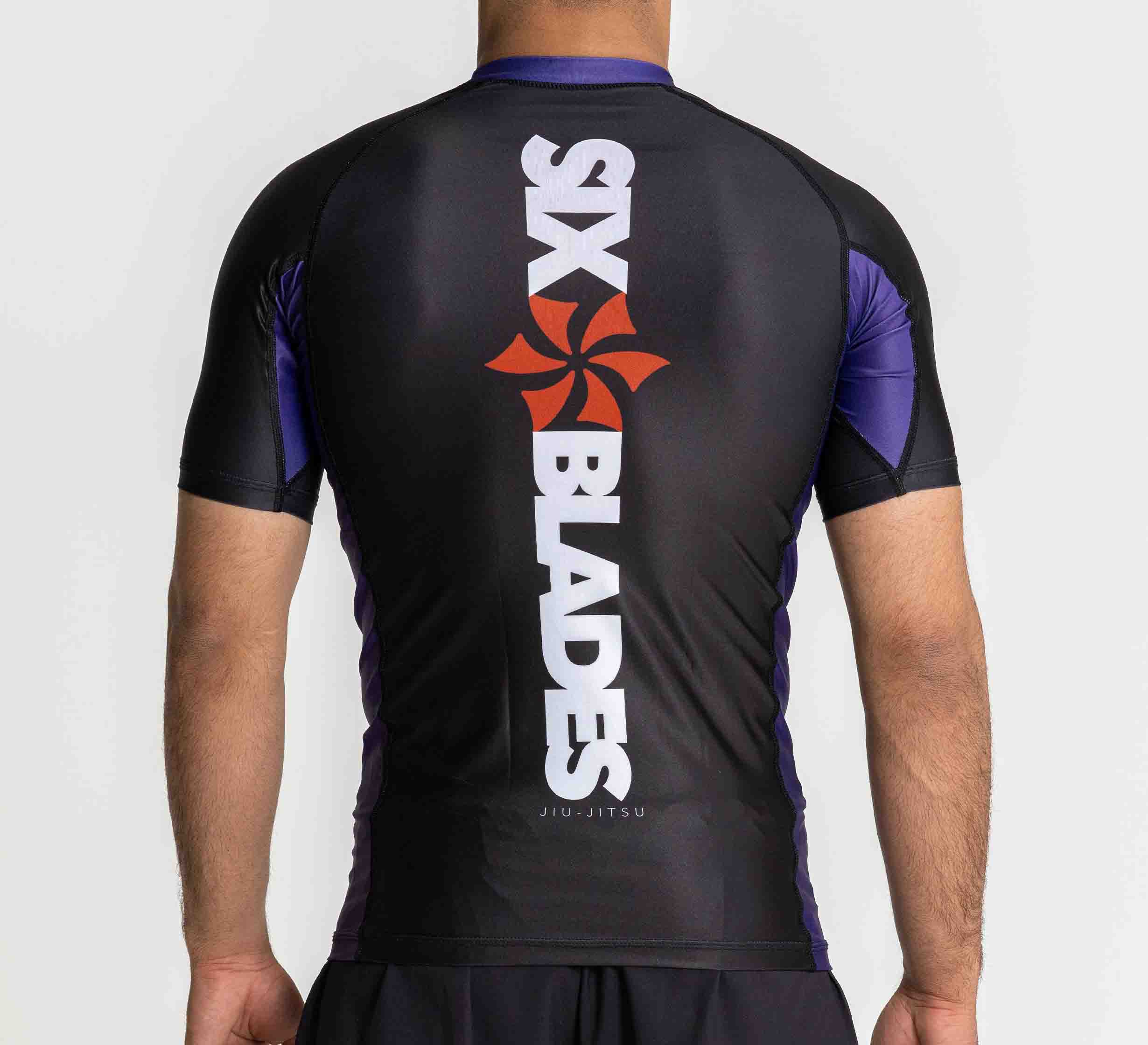A person wearing the Six Blades Short Sleeve Rashguard Purple with a red pinwheel logo and SIX BLADES printed vertically on the back, facing away to show their back, arms, and part of their shorts.