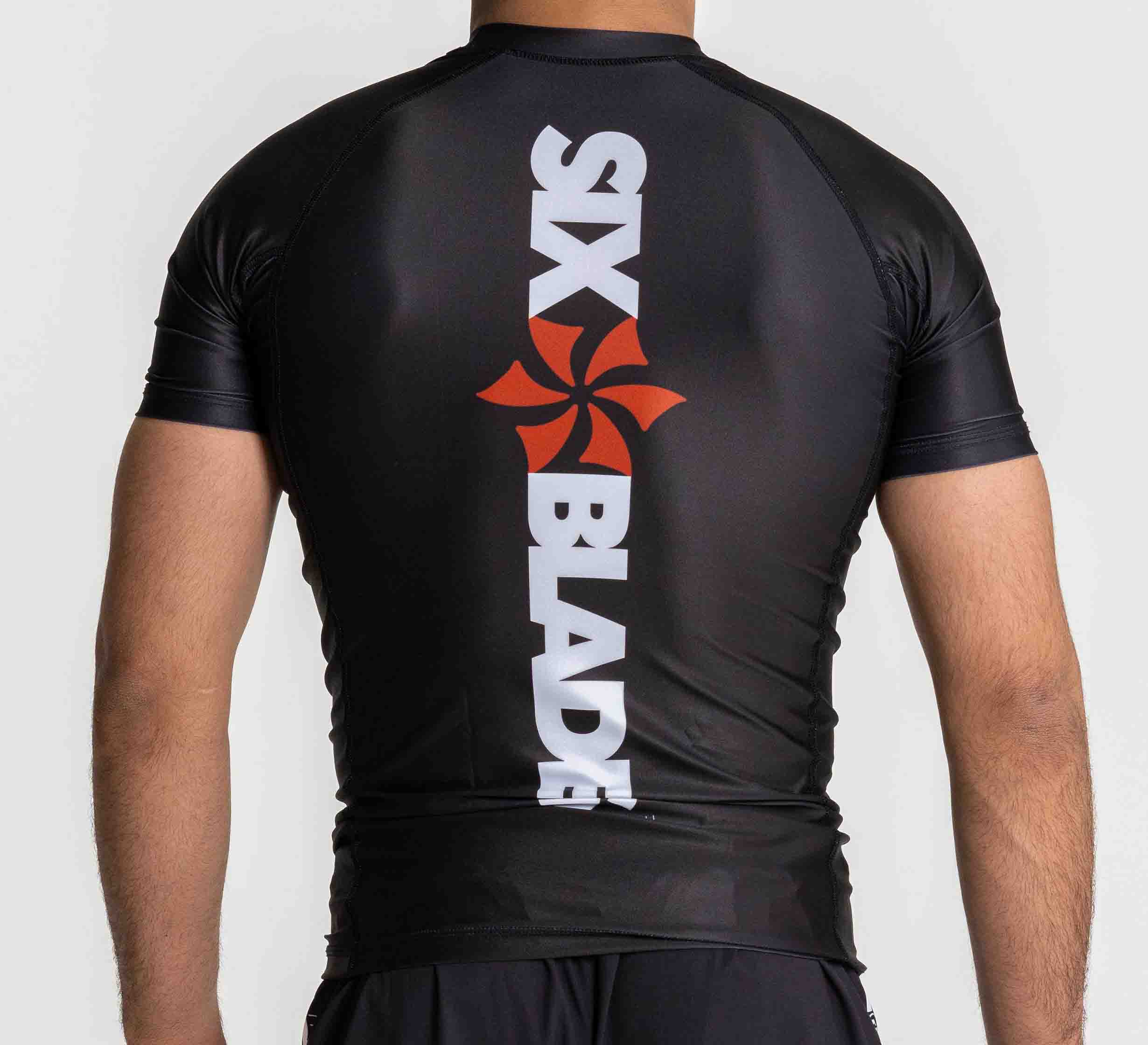 A person is seen from the back wearing the Six Blades Short Sleeve Rashguard Black, featuring a white SIX BLADE logo and a red stylized blade graphic running vertically down the spine.