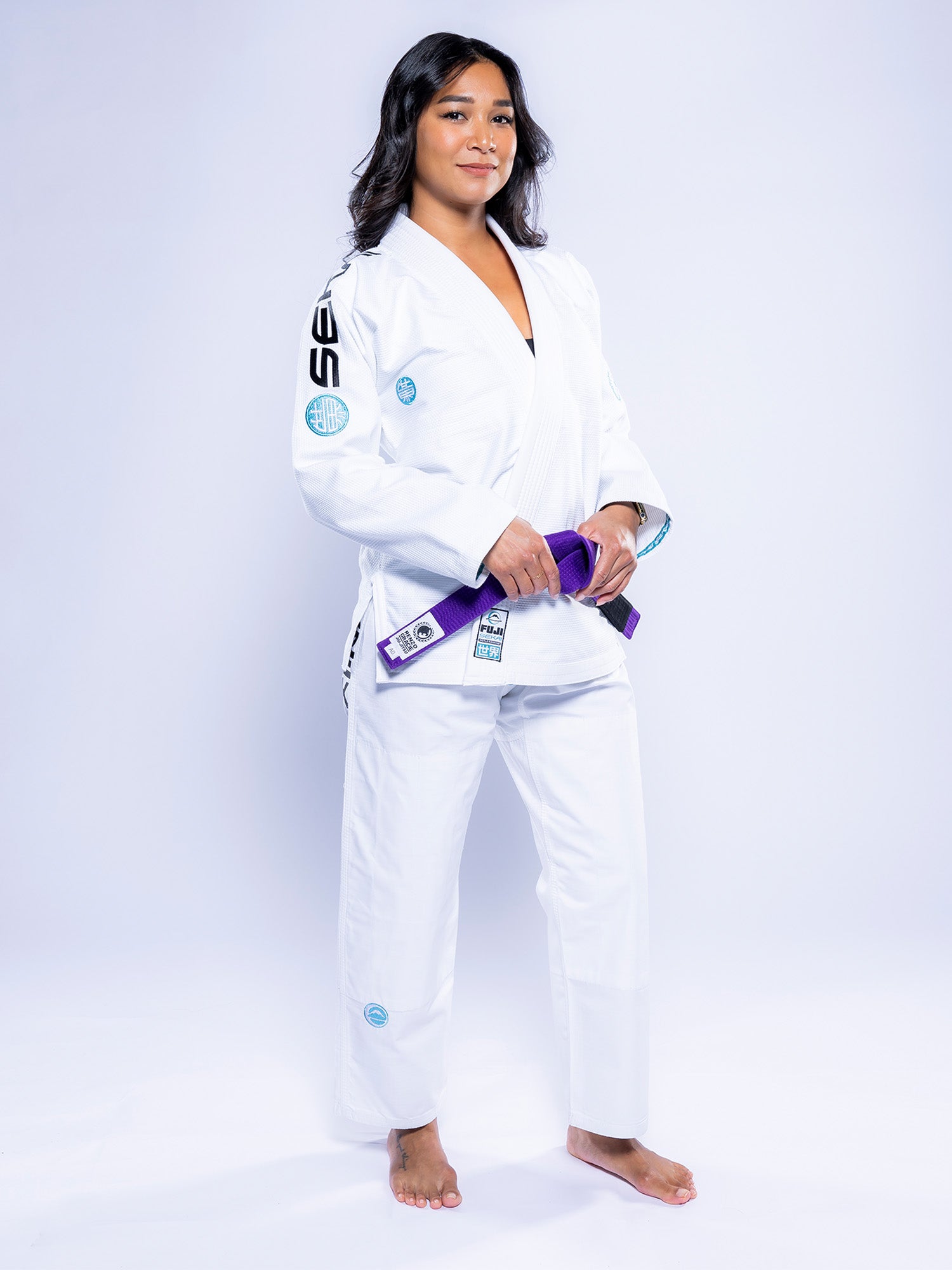 A woman stands barefoot against a white background, confidently holding a purple belt while wearing the Womens SEKAI 3.0 BJJ Gi White Teal with patches.