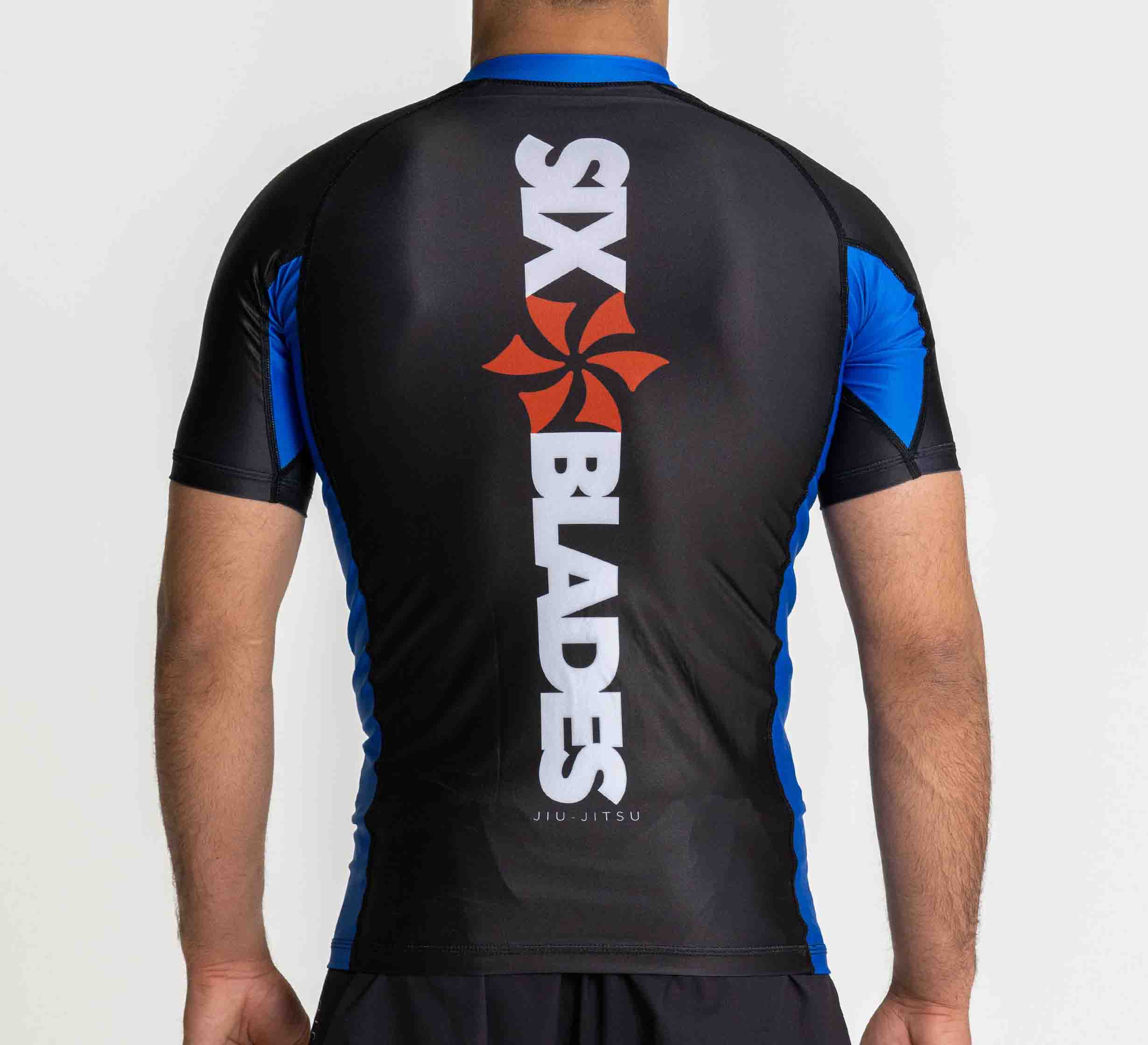 A person wears the Six Blades Short Sleeve Rashguard Blue, featuring black and blue colors with "SIX BLADES JIU-JITSU" and a vertical red blade logo on the back.