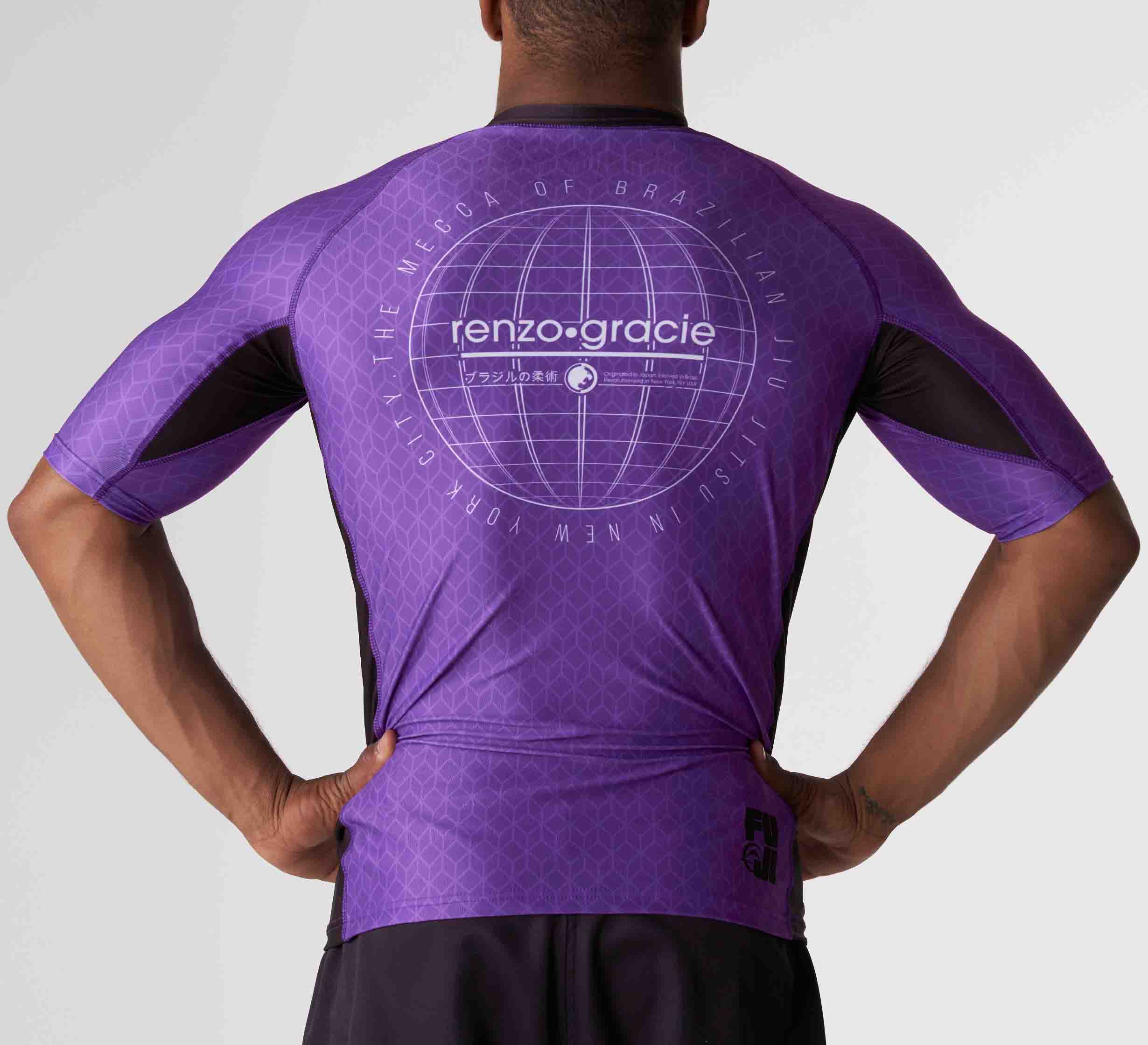A person wearing the Renzo Gracie Mecca Ranked Short Sleeve Rashguard in purple is seen from behind, showcasing the globe graphic and text on the back, hands resting on their hips.