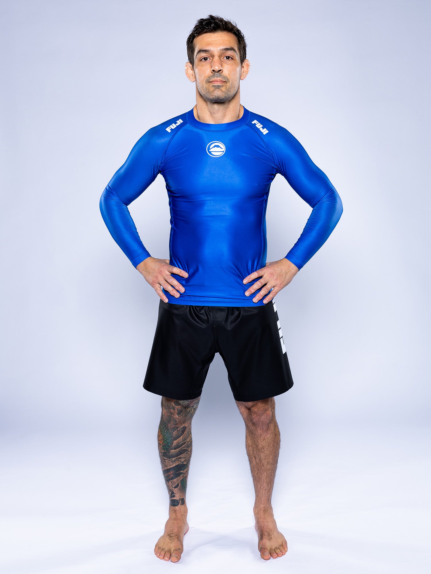 A man with a leg tattoo stands barefoot on a white background, hands on hips, wearing the Baseline 2.0 Long Sleeve Rashguard Blue and black shorts.
