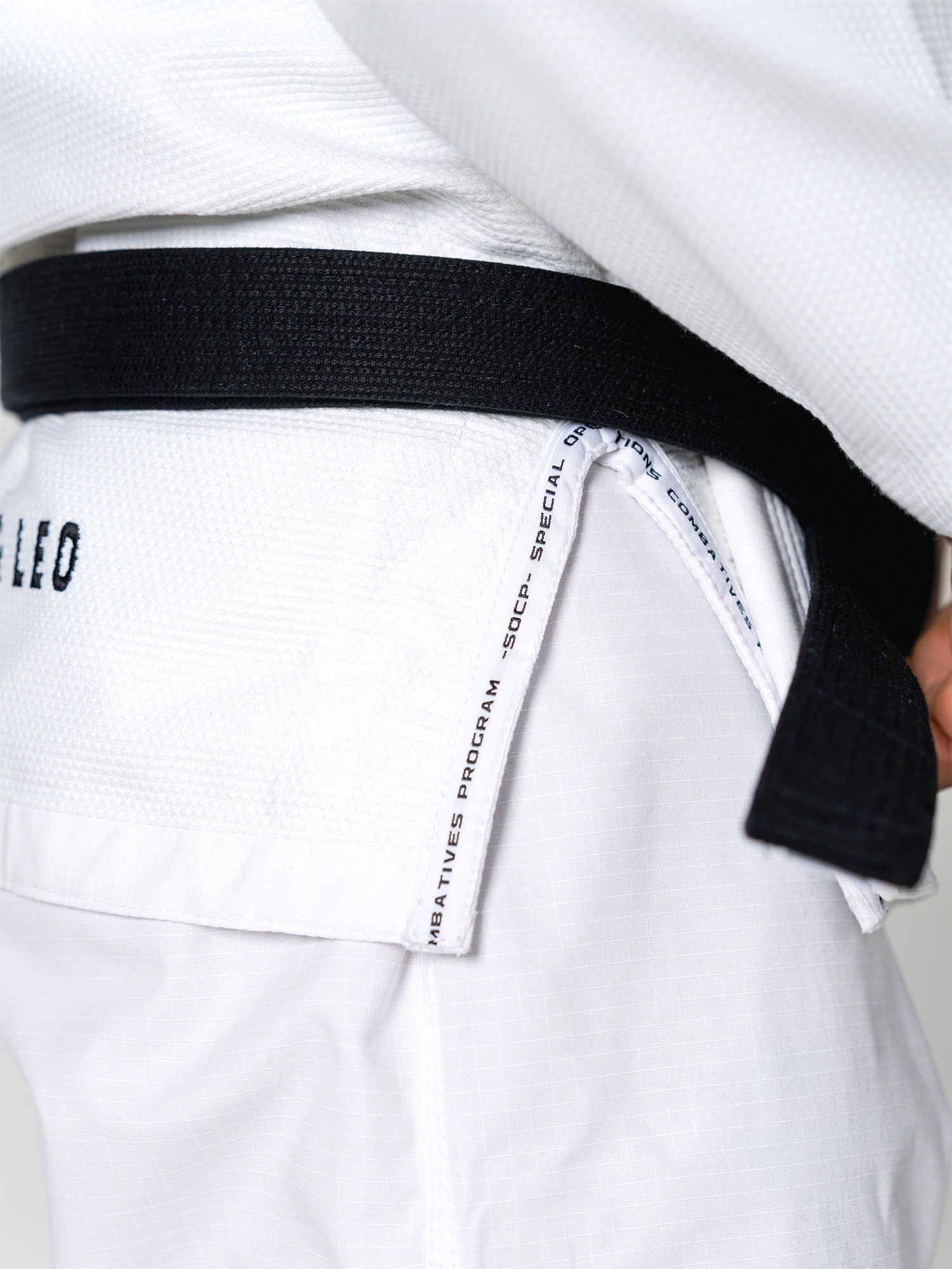 Close-up of a person wearing the S.O.C.P. Sekai BJJ Gi 2.0 White with a black belt. "SOCP SPECIAL COMBATIVES PROGRAM" is visible on the uniform as their hand rests near the belt knot.