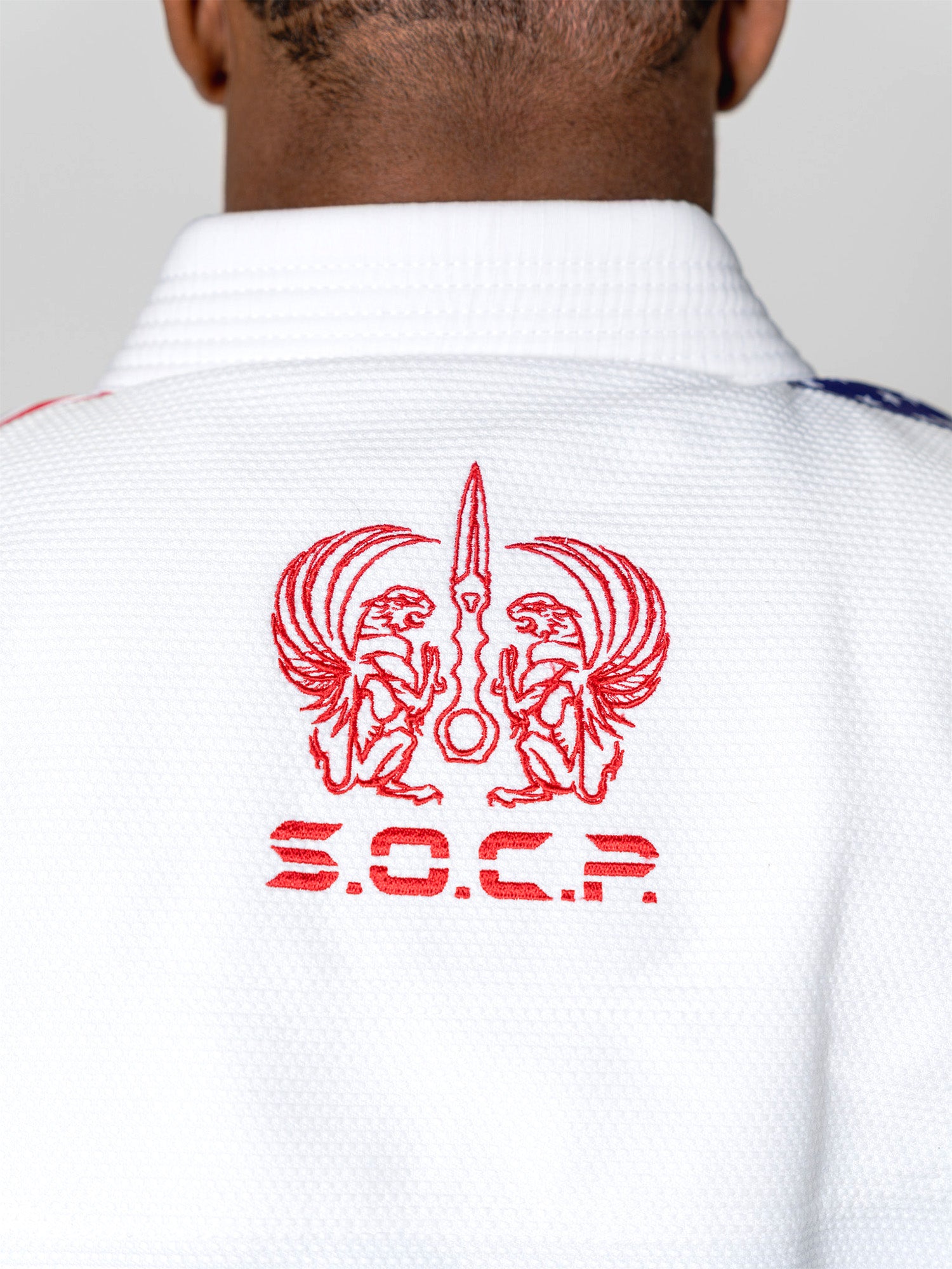A close-up of the S.O.C.P. Sekai BJJ Gi 2.0 White shows its textured fabric, red embroidered twin winged creatures with a sword, and “S.O.C.P.” stitched beneath on the back.