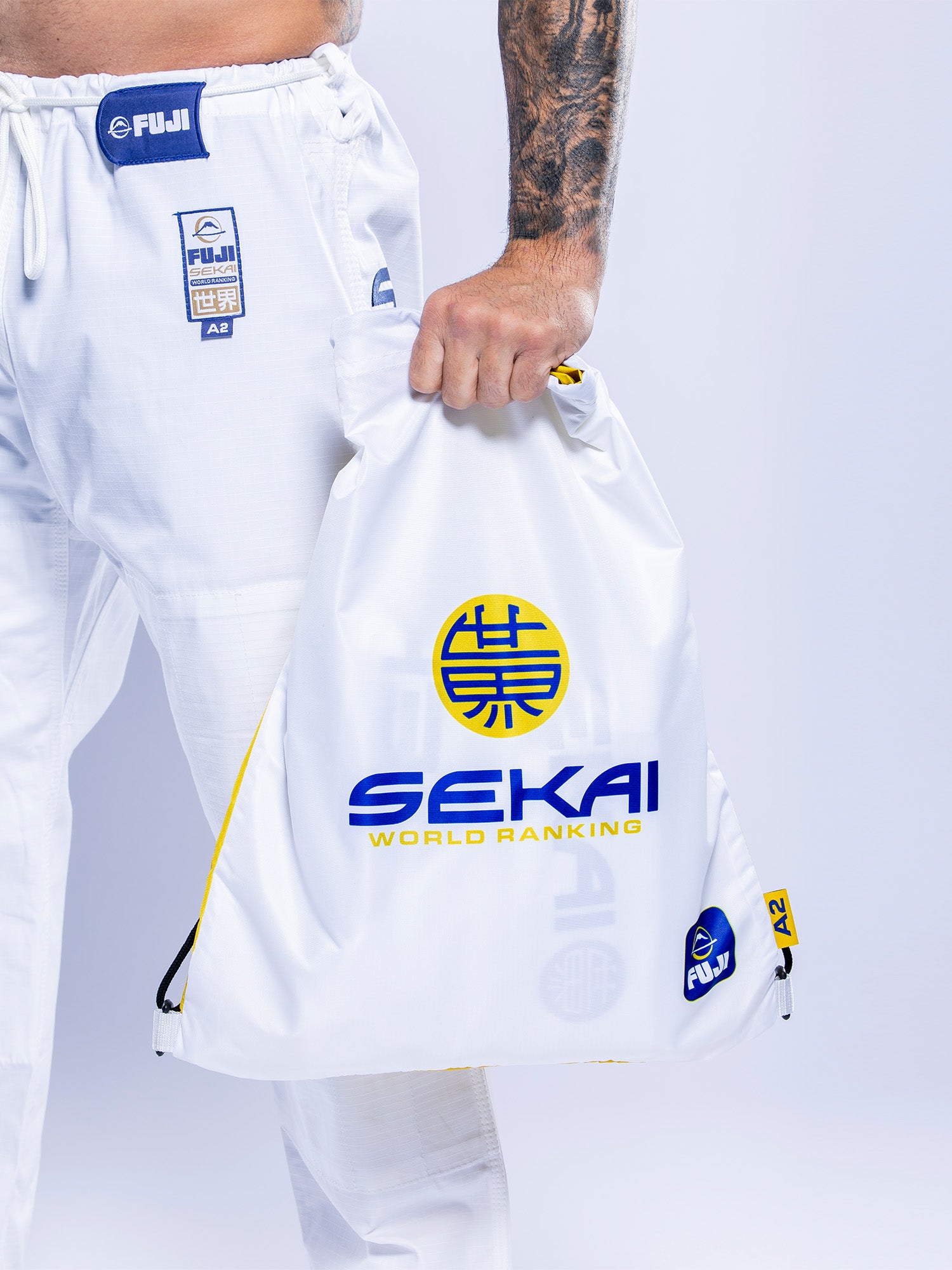 A person in white SEKAI 3.0 BJJ Gi (White Navy) holds a white drawstring bag labeled SEKAI WORLD RANKING with blue, yellow accents, and Japanese characters against a light background.
