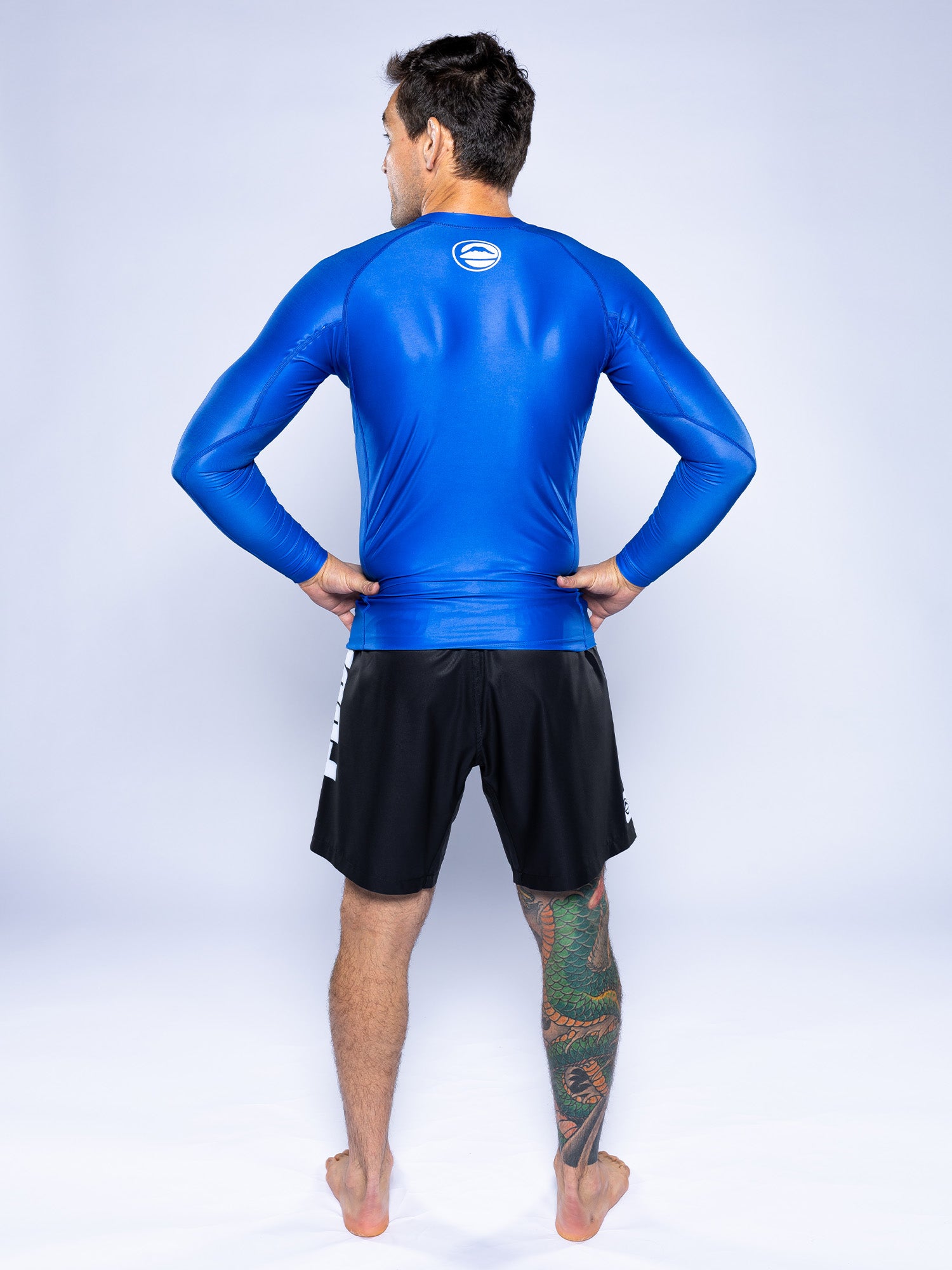 A man stands barefoot, facing away with hands on hips, wearing the Baseline 2.0 Long Sleeve Rashguard Blue and black shorts. His right leg shows a colorful tattoo. The background is plain and light-colored.