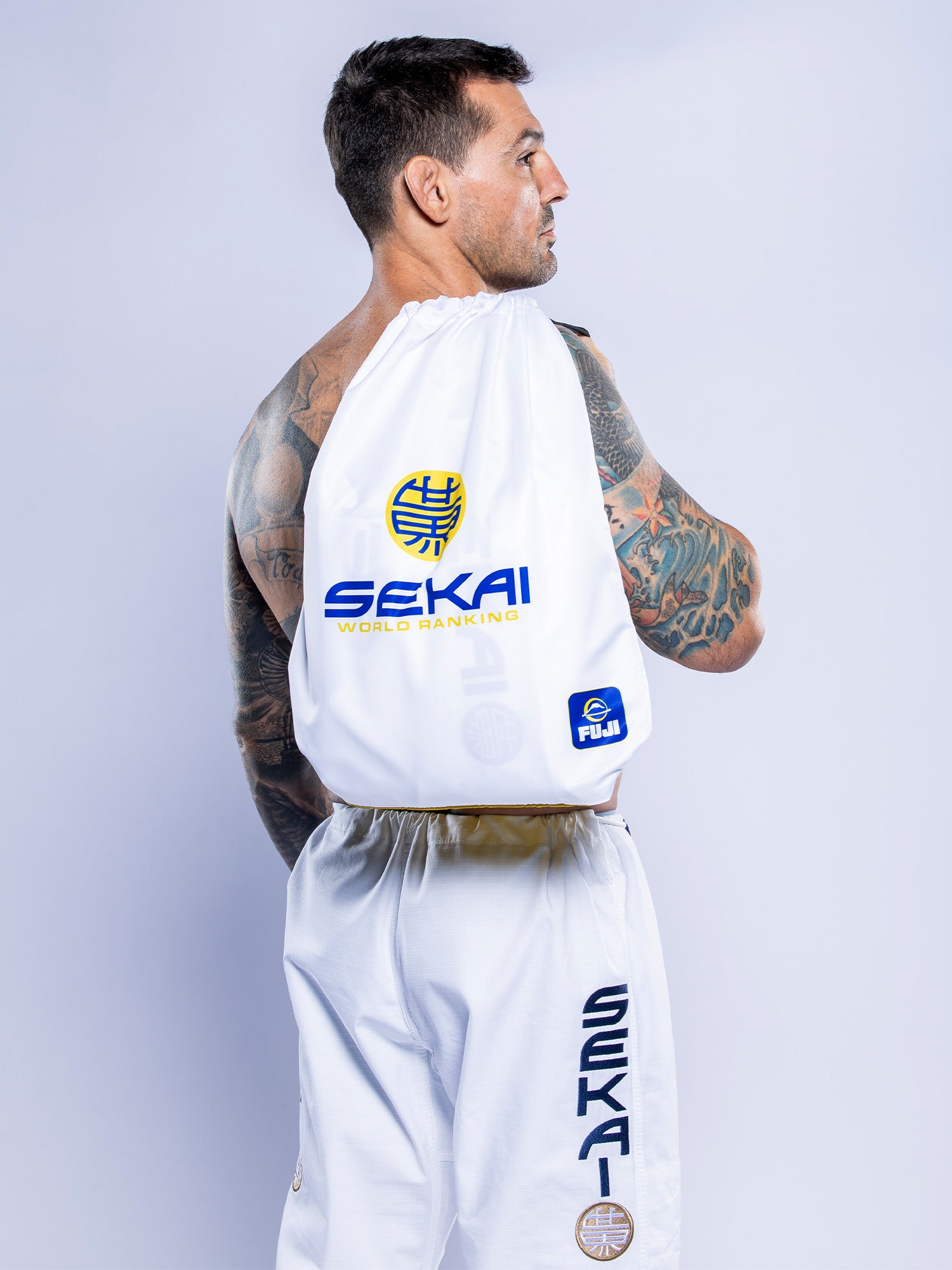 A man with tattooed arms and short dark hair stands sideways wearing the SEKAI 3.0 BJJ Gi White Navy and a white drawstring bag with SEKAI World Ranking and sponsor logos on his back.