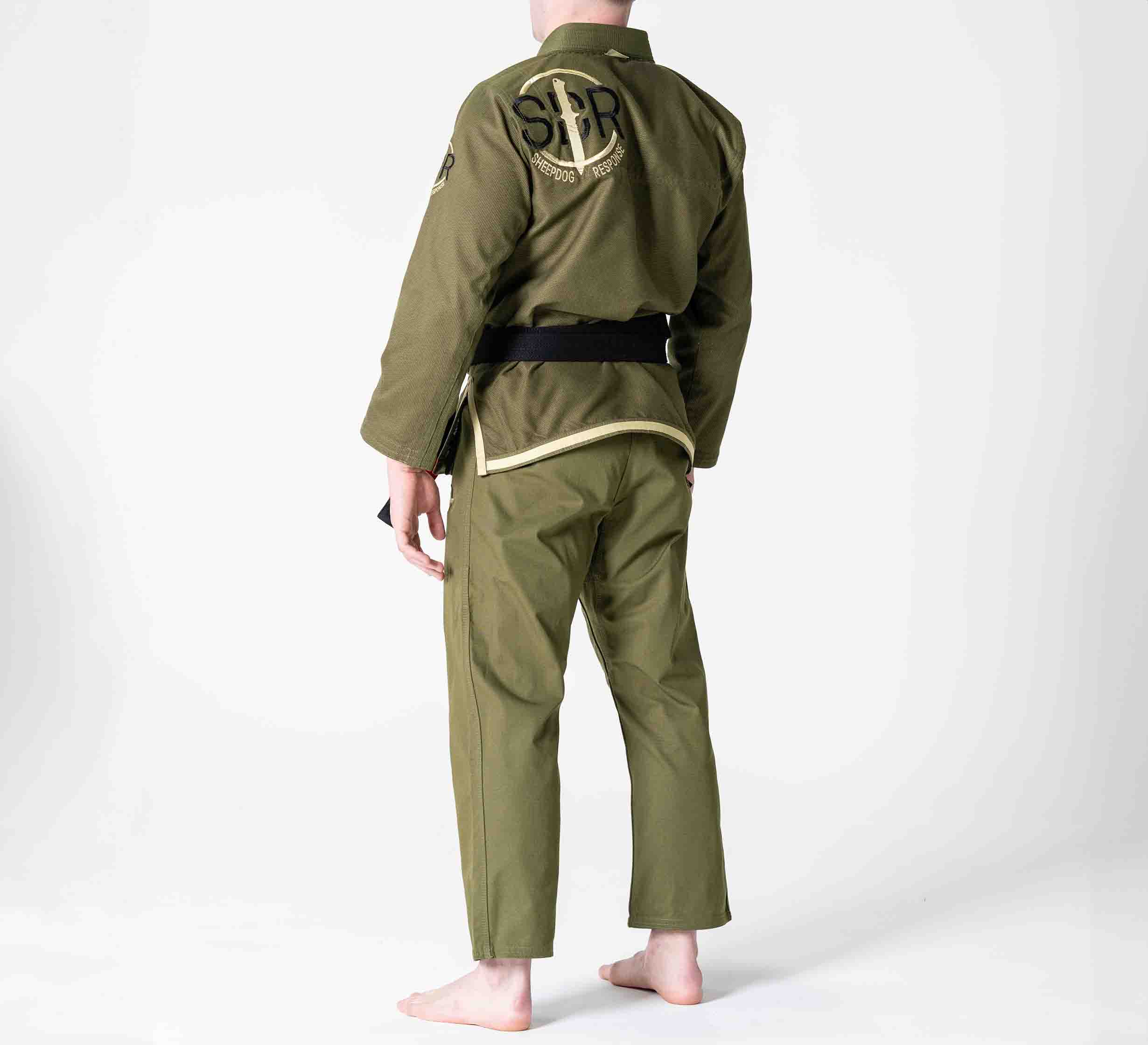 A person stands barefoot, facing away, wearing the Suparaito BJJ Gi Sheepdog Response Edition Military Green with a black belt. The back displays embroidered letters and symbols. The background is plain white.