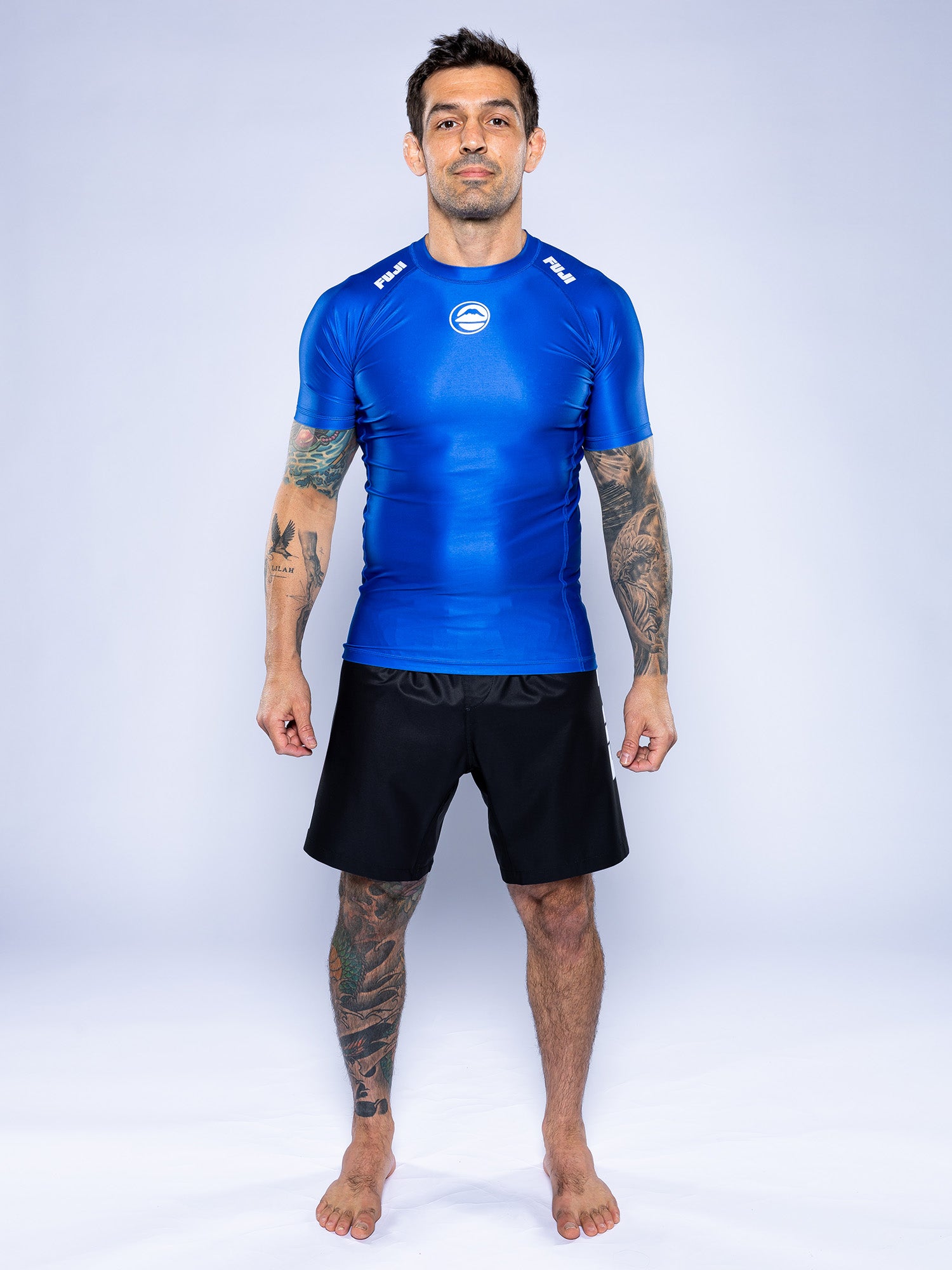 A tattooed man stands barefoot on a white backdrop wearing the Baseline 2.0 Short Sleeve Rashguard Blue and black shorts.