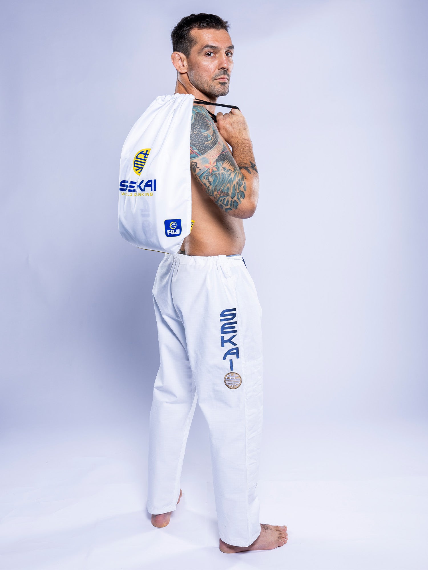 A man with short dark hair and arm tattoos poses in white SEKAI 3.0 BJJ Gi pants, holding the matching white drawstring bag over his shoulder. Both feature navy SEKAI branding and various logos.