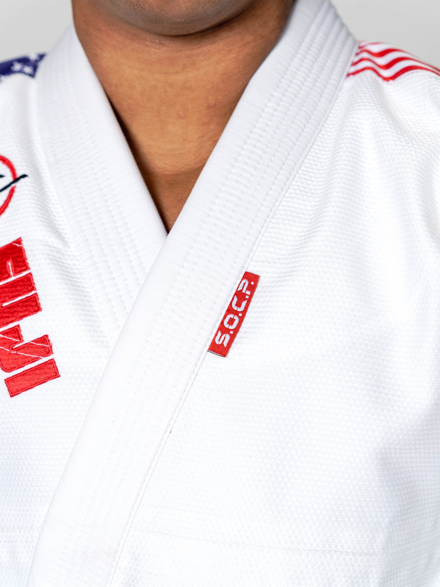 A close-up shows someone wearing the S.O.C.P. Sekai BJJ Gi 2.0 White with red patches and an American flag on the collar. The person's face is partially out of frame.