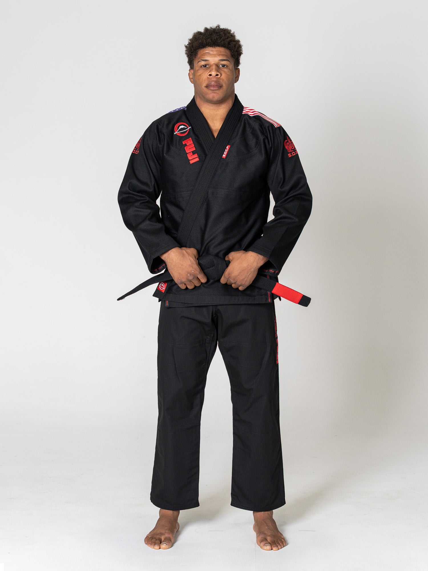 A person stands barefoot on a white background, wearing the S.O.C.P. Sekai BJJ Gi 2.0 Black with red patches and a black belt featuring a red stripe, hands holding the belt in front.