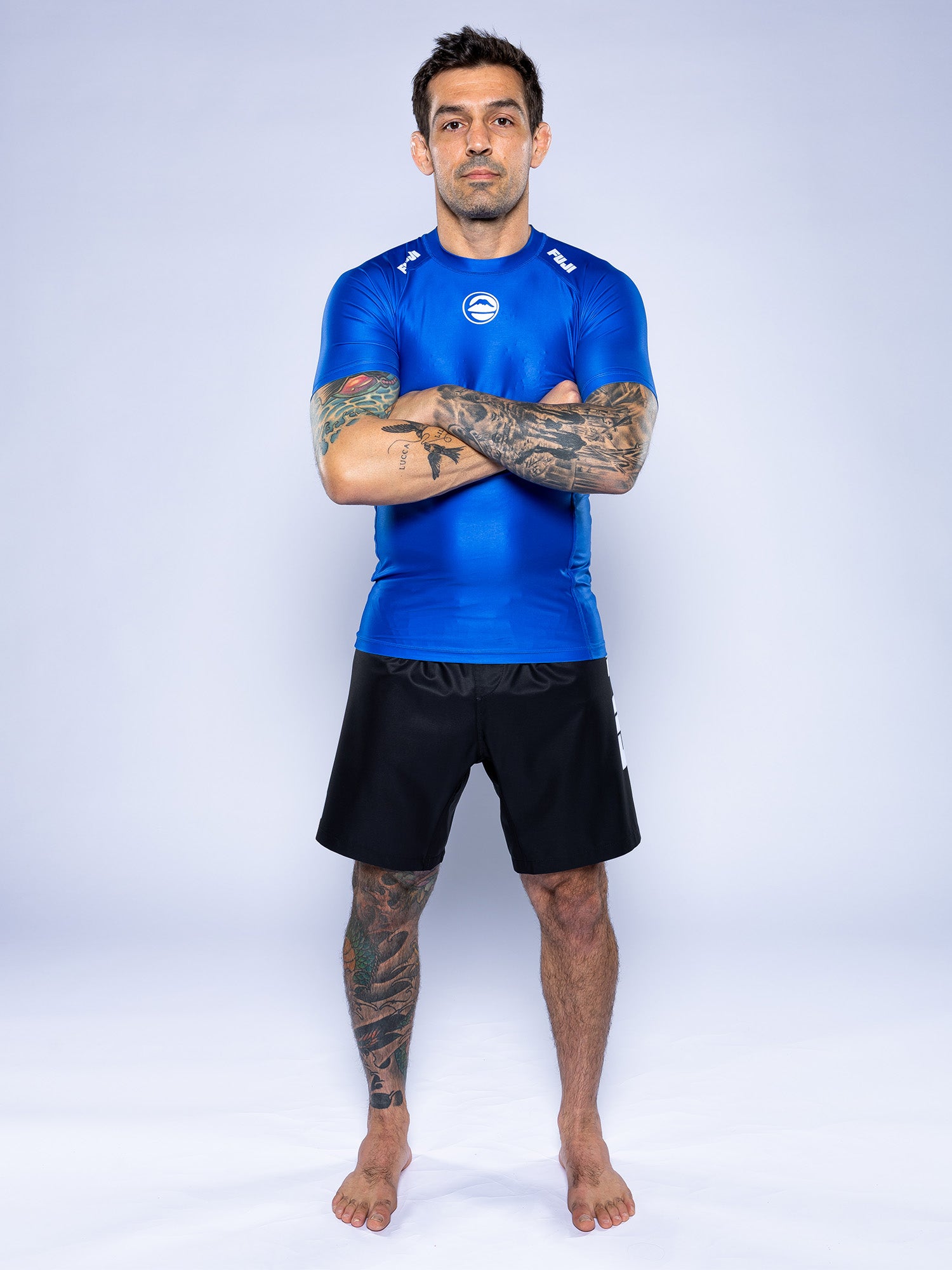 A man with short dark hair and tattoos stands barefoot on a white background, arms crossed, wearing the Baseline 2.0 Short Sleeve Rashguard Blue and black shorts.