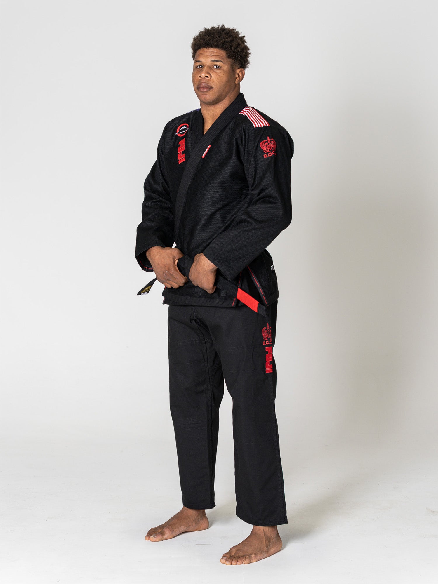 A person stands barefoot on a white background, wearing the S.O.C.P. Sekai BJJ Gi 2.0 Black with red patches and details, hands adjusting the belt while looking toward the camera.