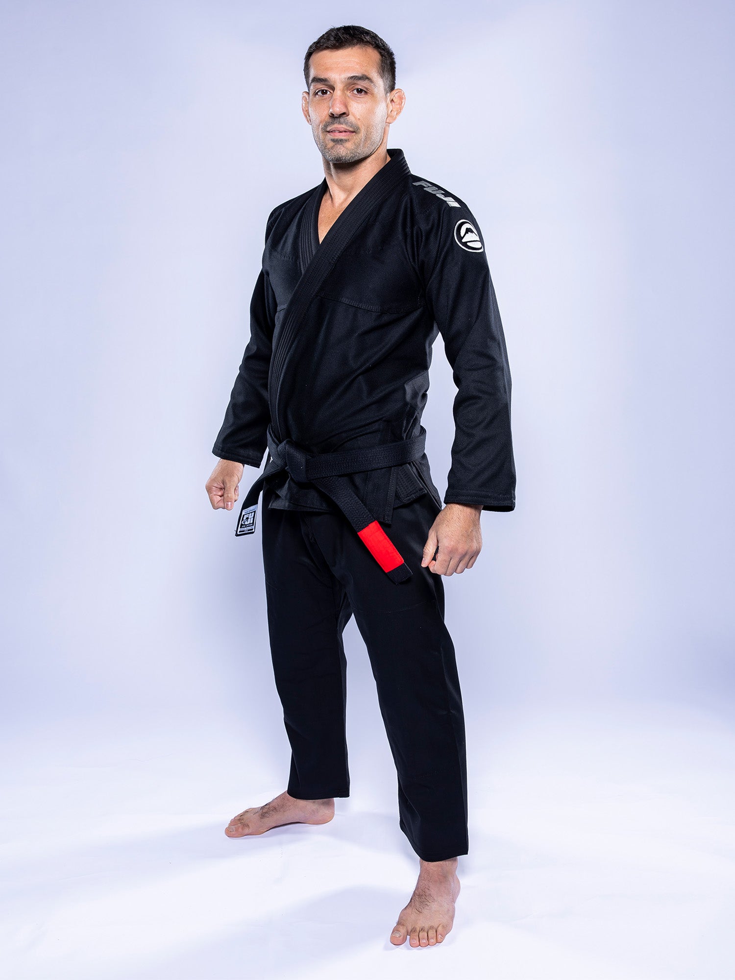 A man stands barefoot on a white background, confidently facing the camera with arms at his sides, wearing the Baseline 2.0 BJJ Gi Black Grey and a black belt featuring a red bar.