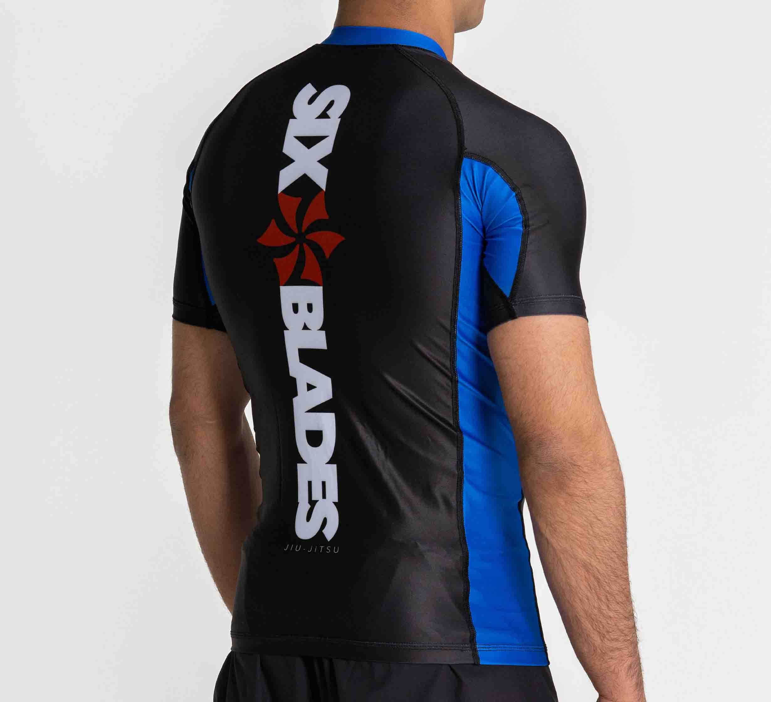A person stands with their back to the camera, wearing the Six Blades Short Sleeve Rashguard Blue featuring "SIX BLADES" and a red pinwheel logo on the back.