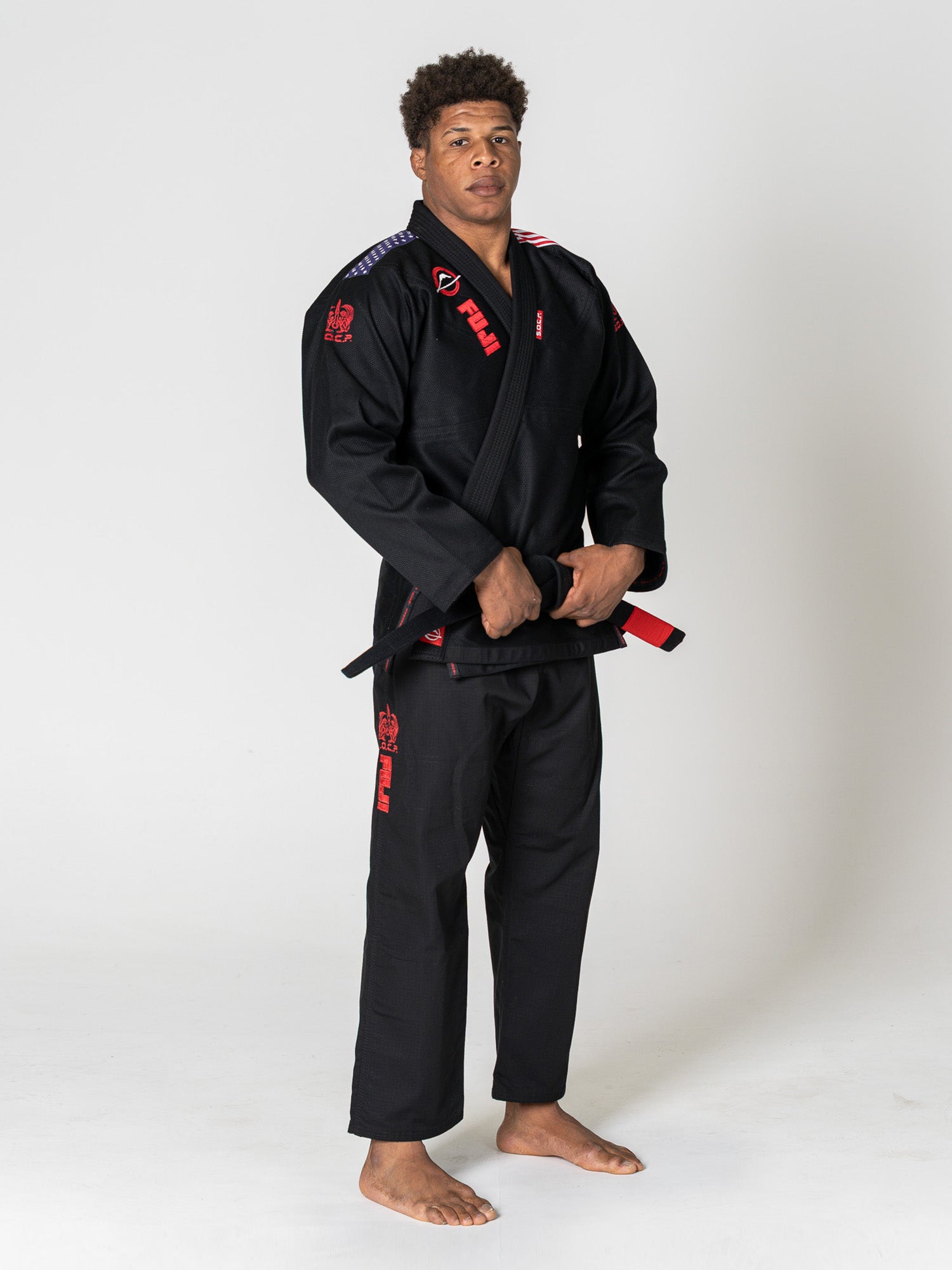 A man stands barefoot on a white background, wearing the S.O.C.P. Sekai BJJ Gi 2.0 Black with red patches, holding the ends of his black belt in front of him.