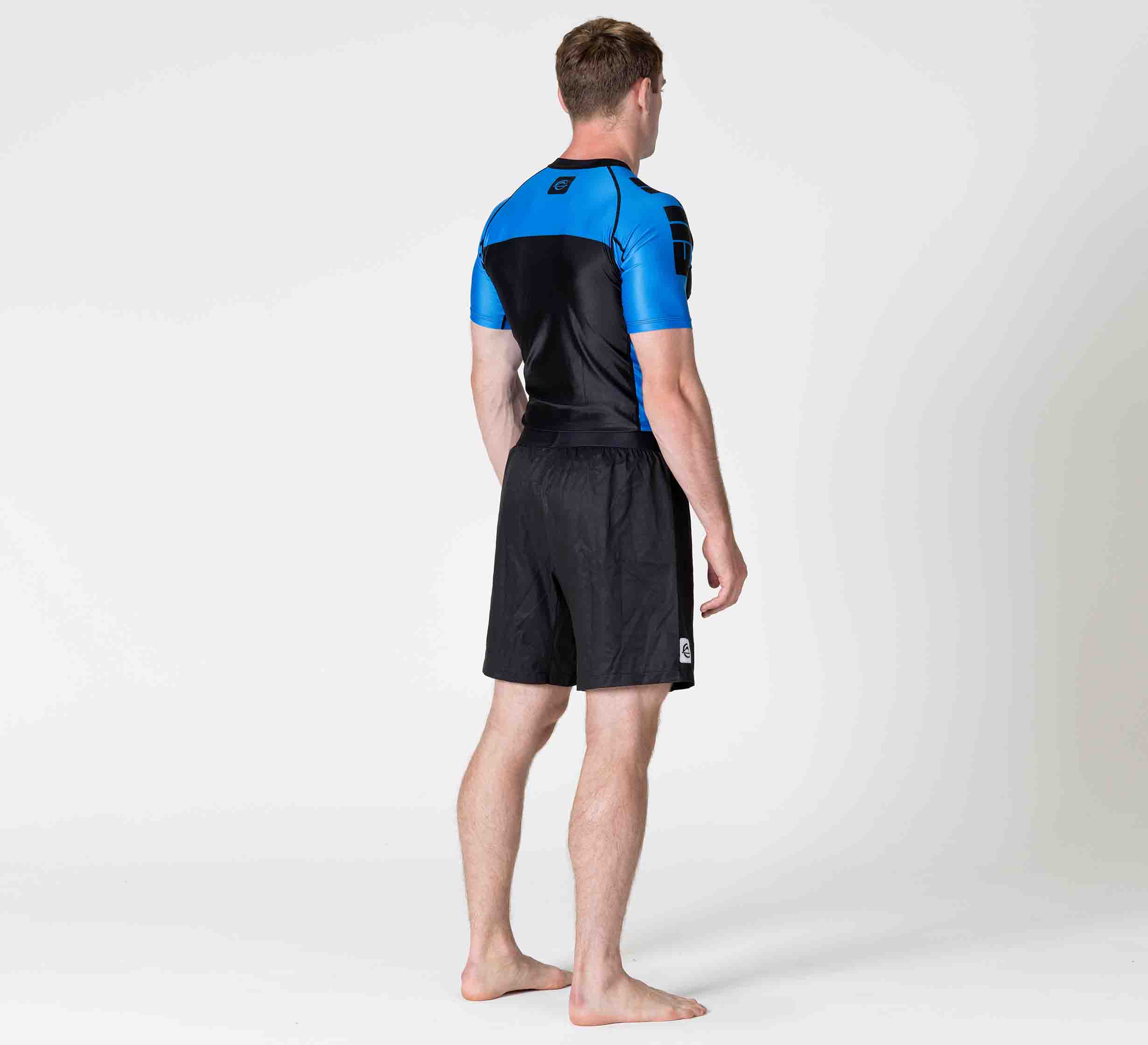 A man stands barefoot on a white background, facing away. He is wearing the Competition Ranked Rashguard Blue and black shorts.