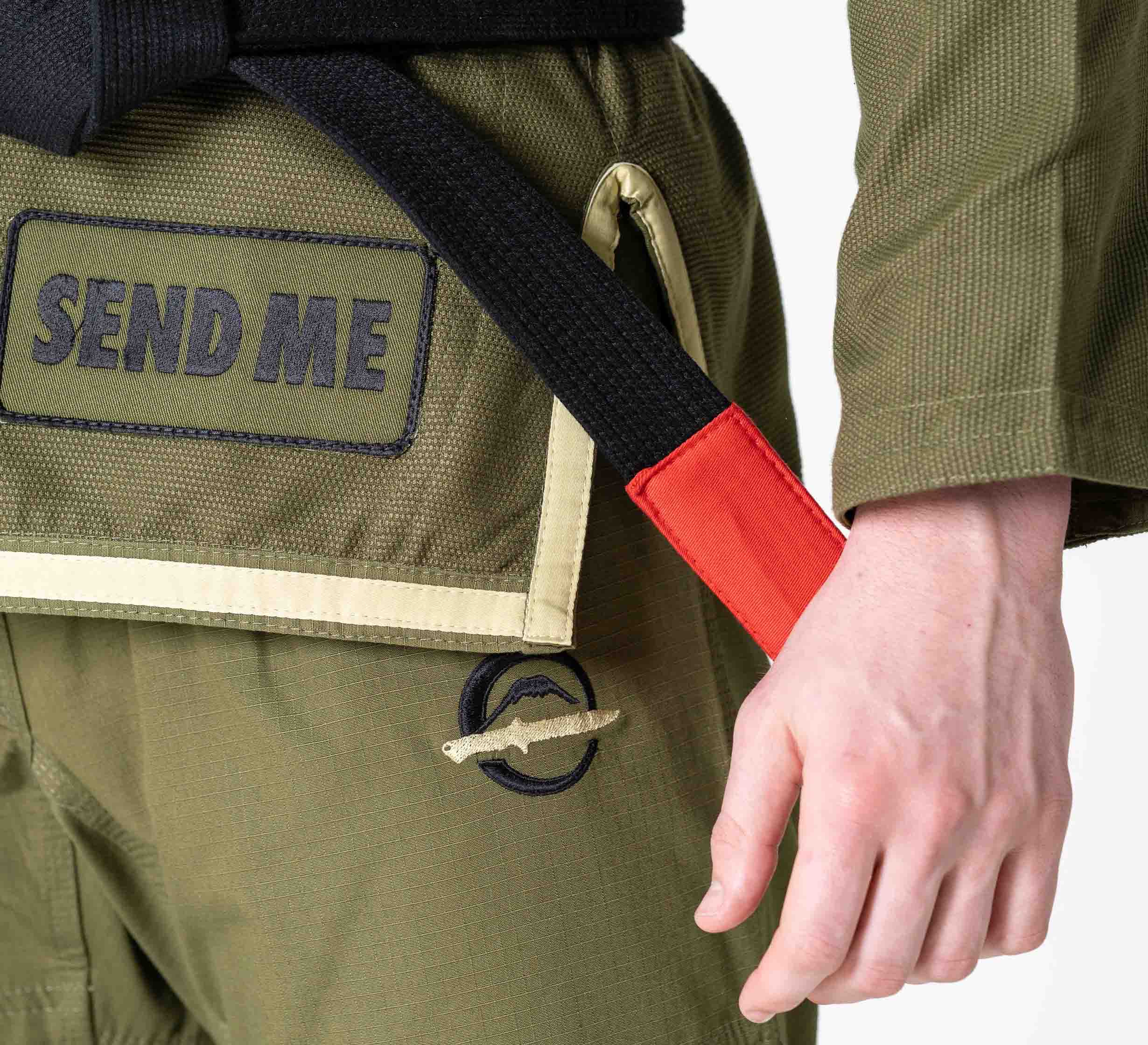 Close-up of a person wearing the Suparaito BJJ Gi Sheepdog Response Edition in military green with a black belt featuring a red tip. The gi displays a SEND ME patch, and the person's right hand rests near the belt.