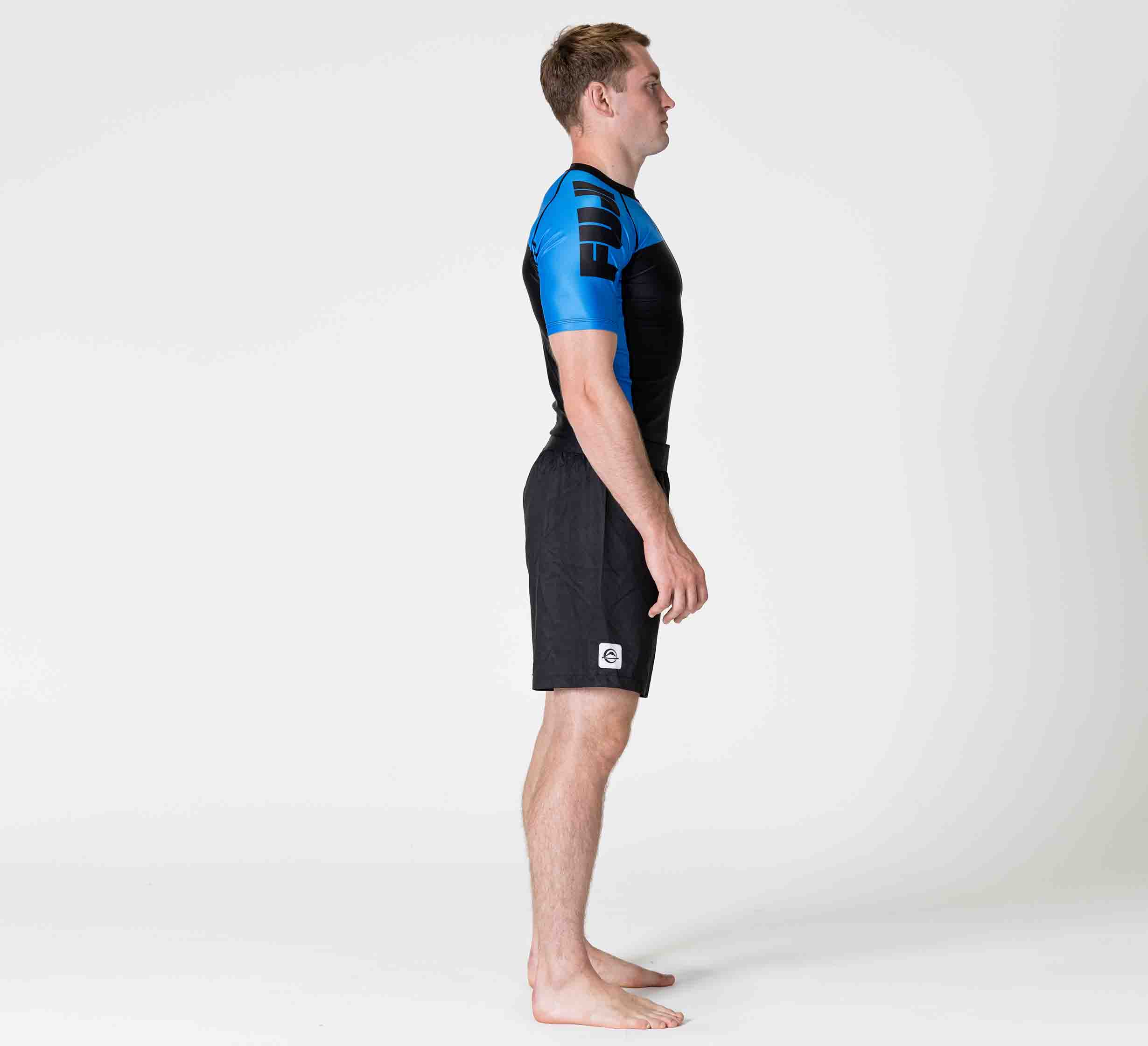 A man stands in profile on a white background, wearing the Competition Ranked Rashguard Blue and black shorts. He is barefoot, looking straight ahead with his arms relaxed at his sides.