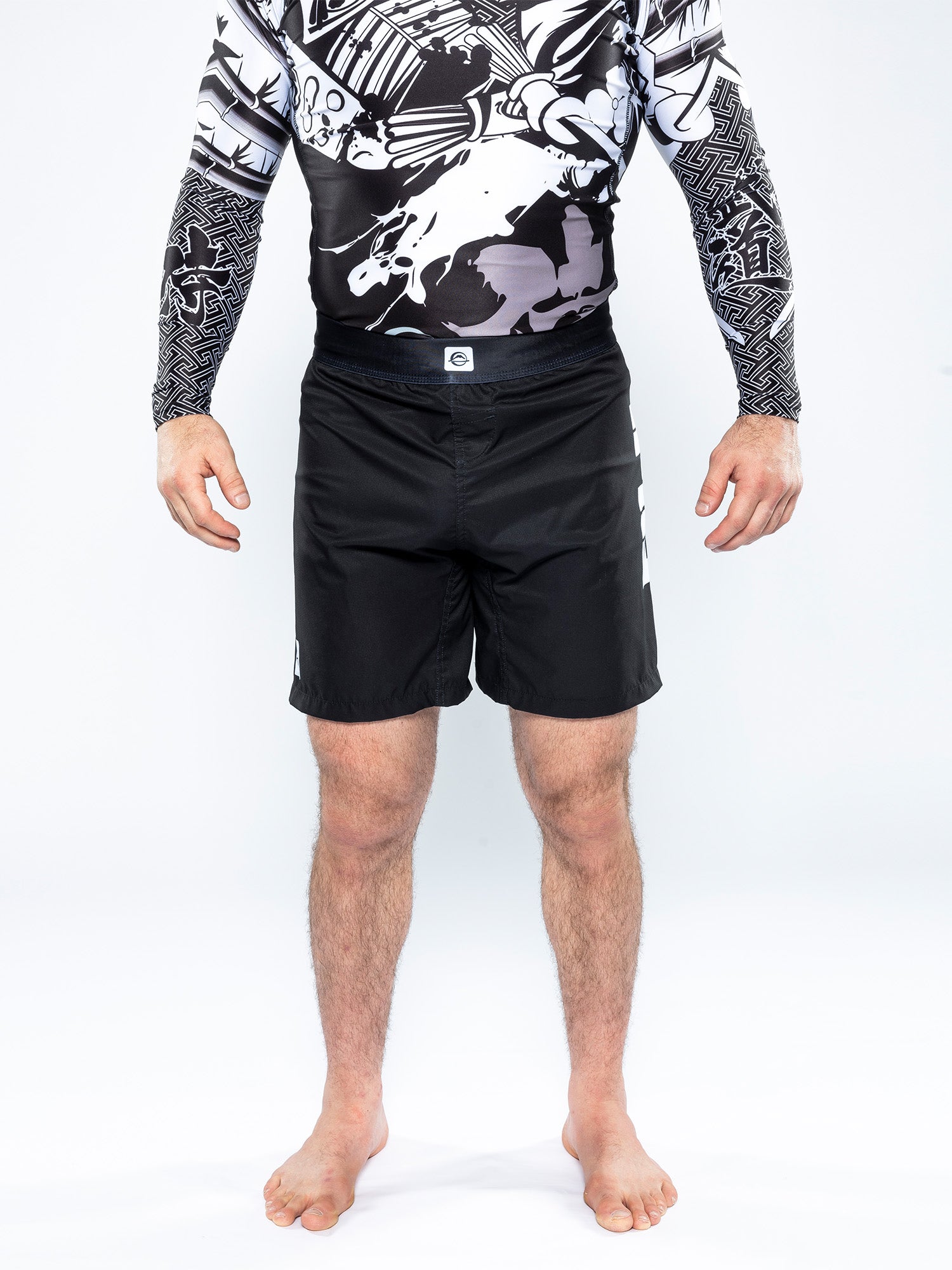 A person stands barefoot wearing Competition Ranked Shorts Black and a long-sleeve black-and-white rash guard, shown from shoulders to feet against a plain white background.