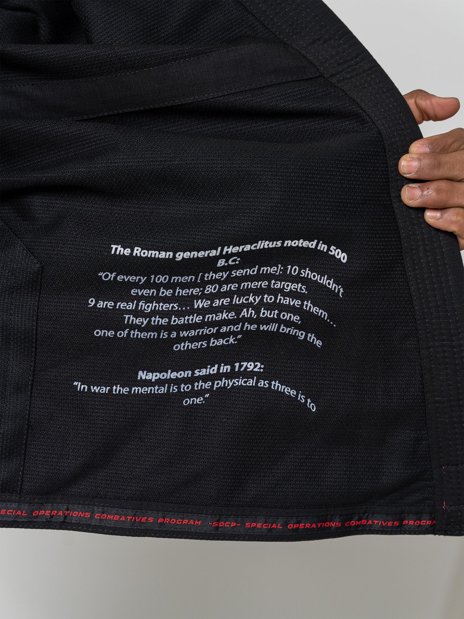 Close-up of the S.O.C.P. Sekai BJJ Gi 2.0 Black showing white warrior quotes from Heraclitus (500 B.C.) and Napoleon (1792) printed on black fabric, with a hand holding the edge and red stitched text partially visible at the bottom.
