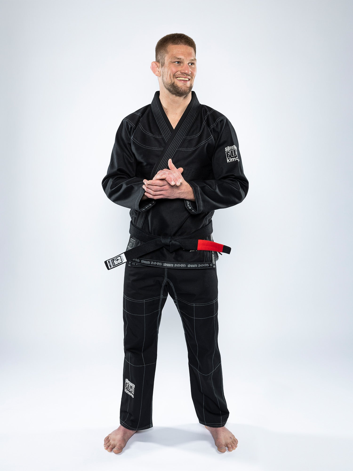 A man in a Suparaito BJJ Gi and black belt stands barefoot on a white background, smiling with his hands together in front of him.