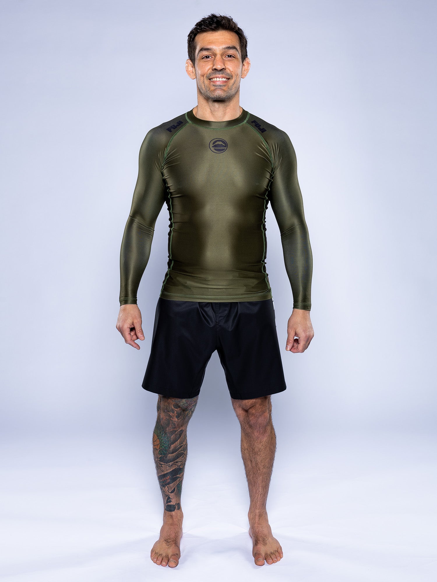 A man stands barefoot against a plain backdrop, smiling. He wears the Baseline 2.0 Long Sleeve Rashguard in Military Green and black shorts, with a tattoo visible on his left leg.