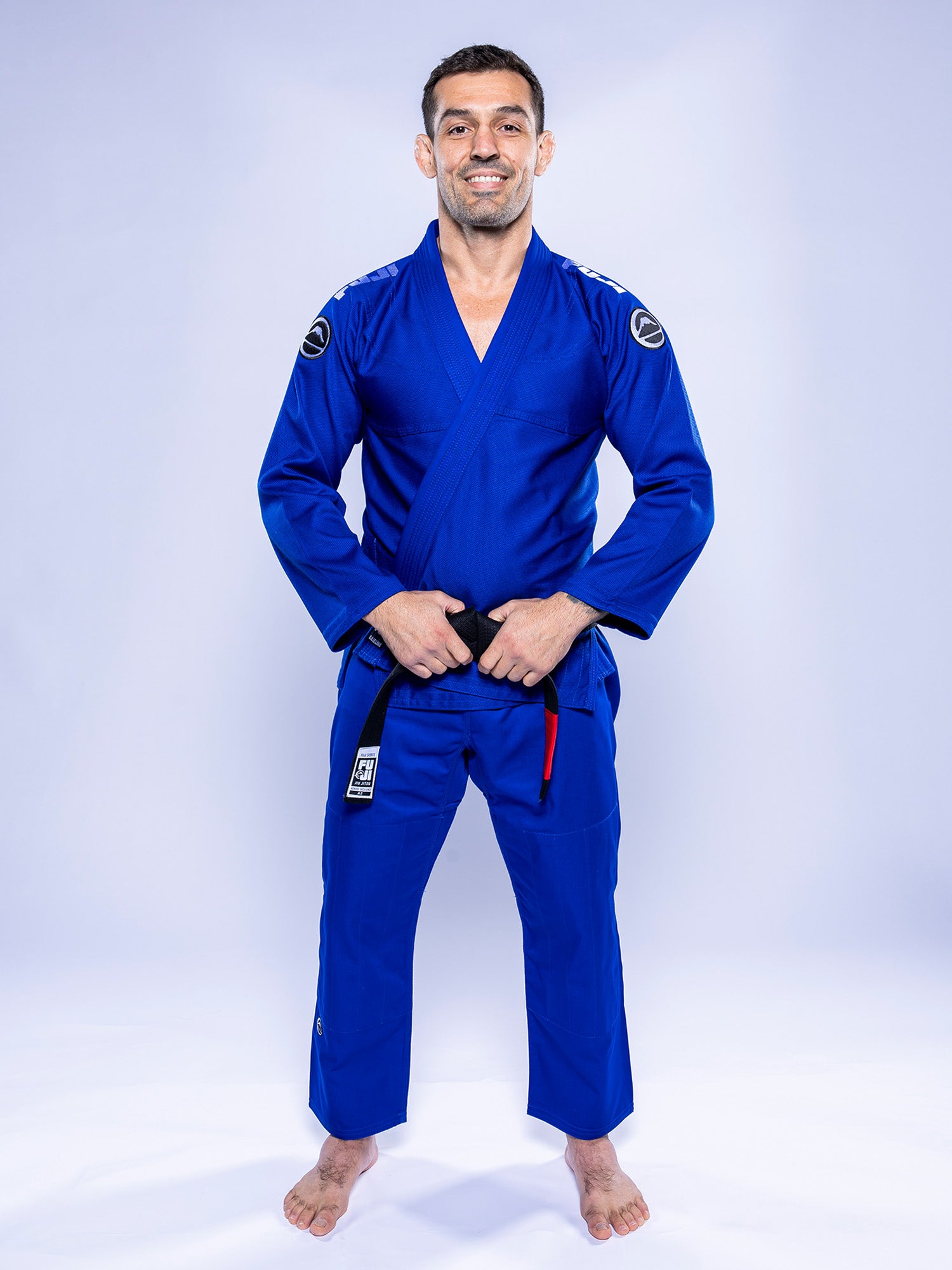 A man stands barefoot on a white background, smiling and holding his belt while wearing the Baseline 2.0 BJJ Gi Blue White and a black belt.