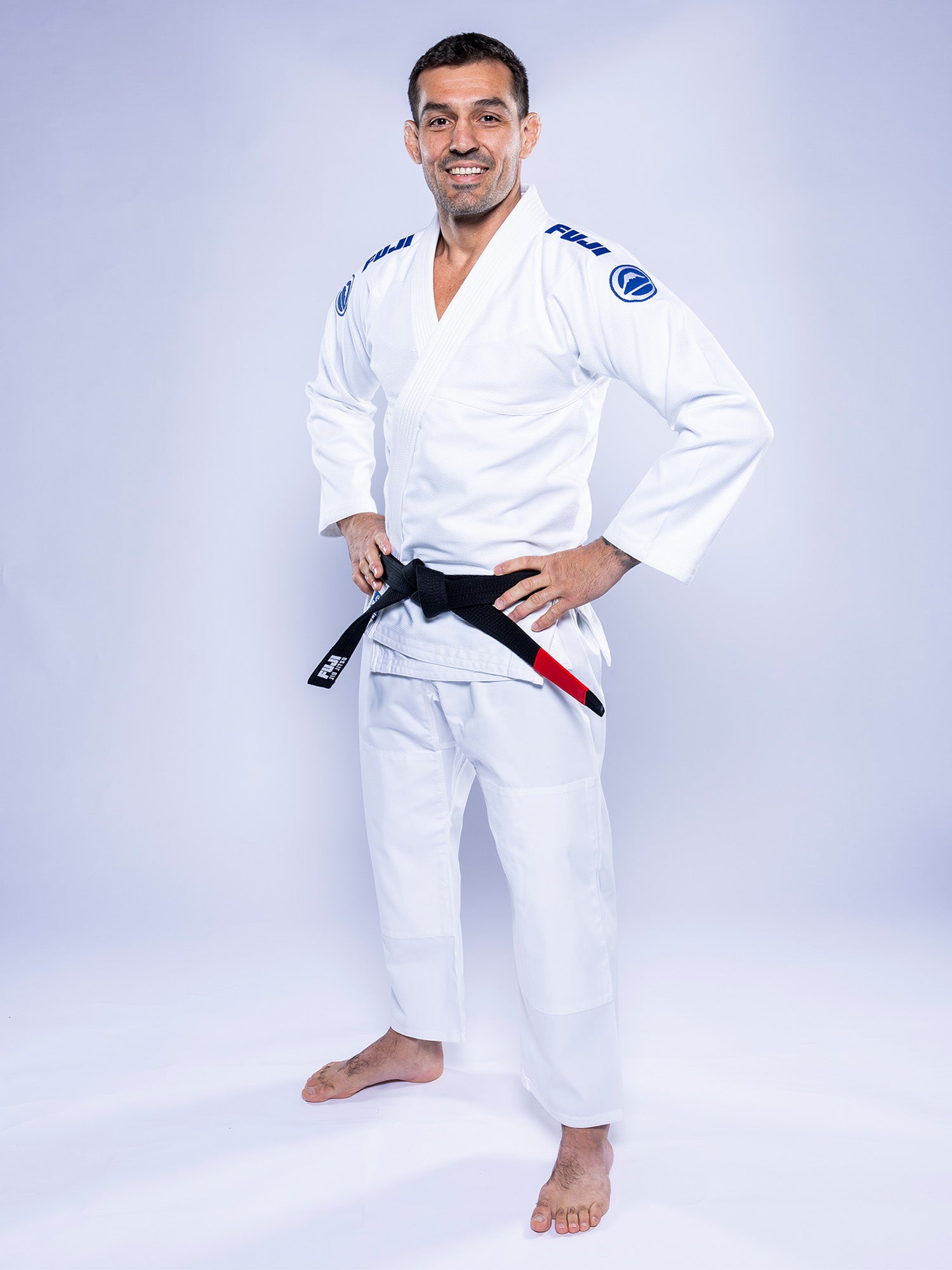 A man wearing the Baseline 2.0 BJJ Gi White Navy and a black belt stands barefoot on a light background, smiling with his hands on his hips.
