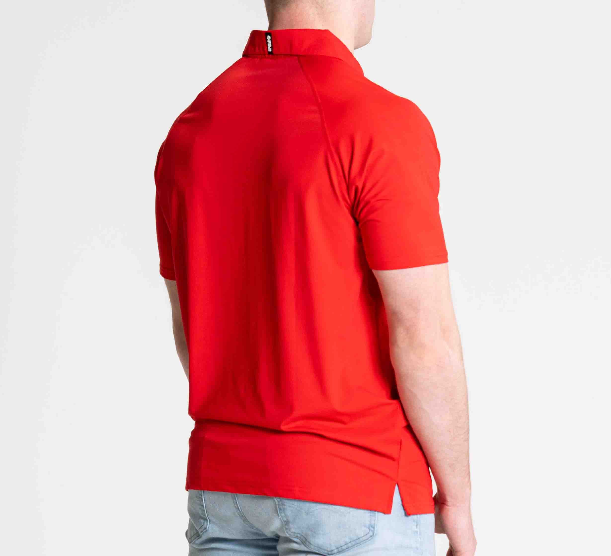 Someone stands facing away from the camera against a plain white background, wearing the FUJI Performance Polo Red and light blue jeans.