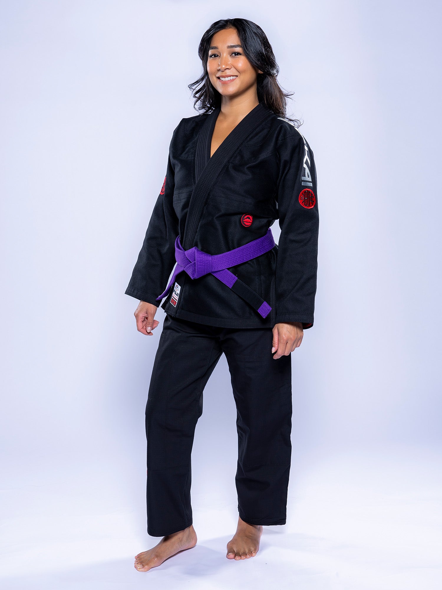 A woman stands barefoot on a light background, smiling and wearing the Womens SEKAI 3.0 BJJ Gi Black Grey with a purple belt tied at her waist.