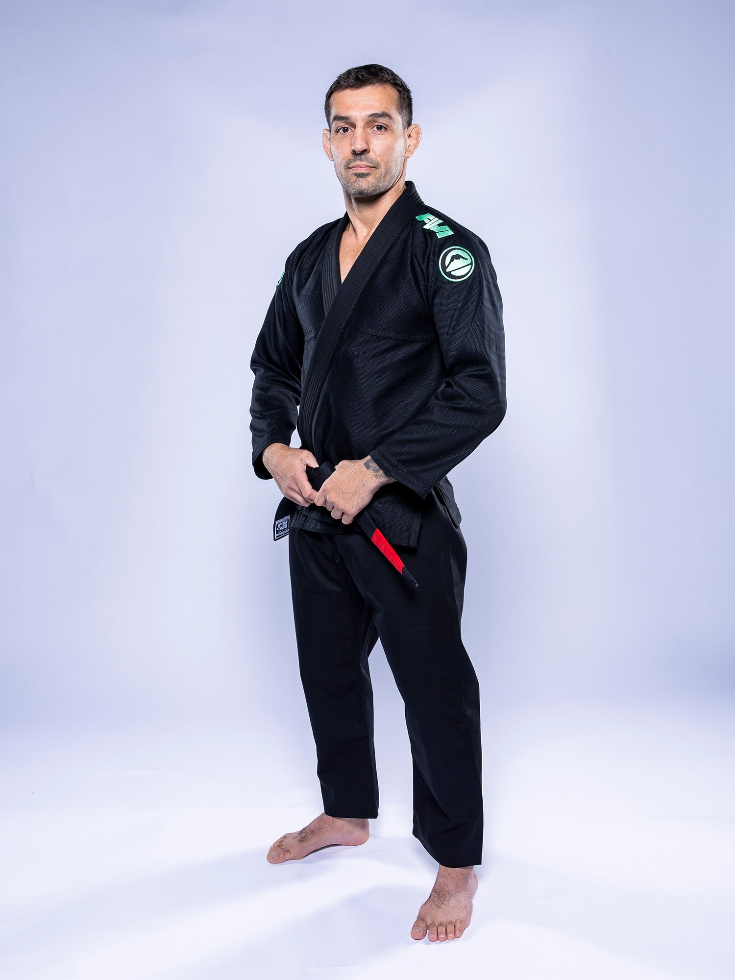 A man stands barefoot on a white surface, wearing the Baseline 2.0 BJJ Gi Black Mint and a red-banded black belt. He faces forward with a neutral expression, holding his belt against a plain light background.