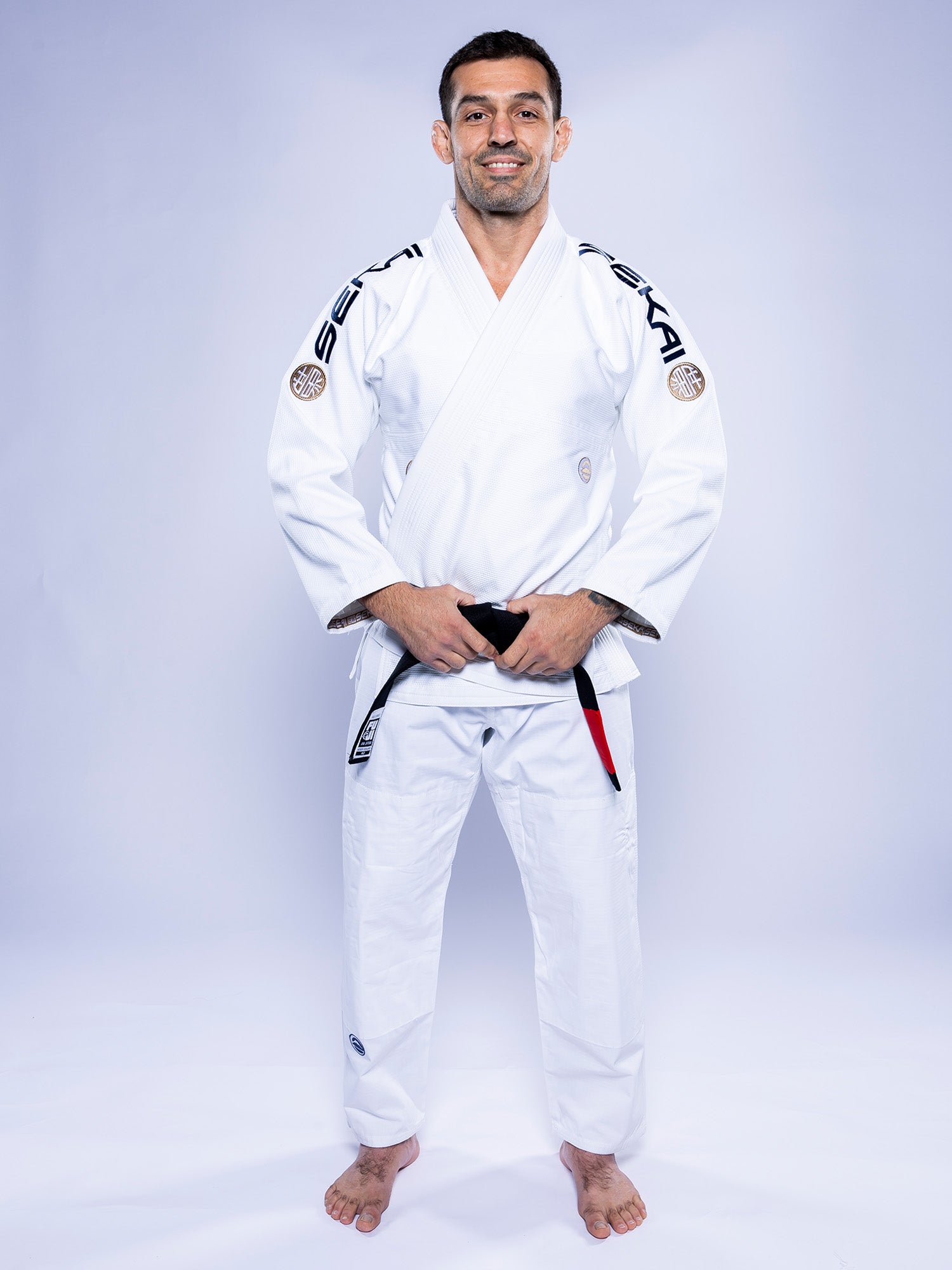 A man wearing the SEKAI 3.0 BJJ Gi White Navy with a black belt stands barefoot on a white background, smiling and facing forward with his hands resting on his belt.
