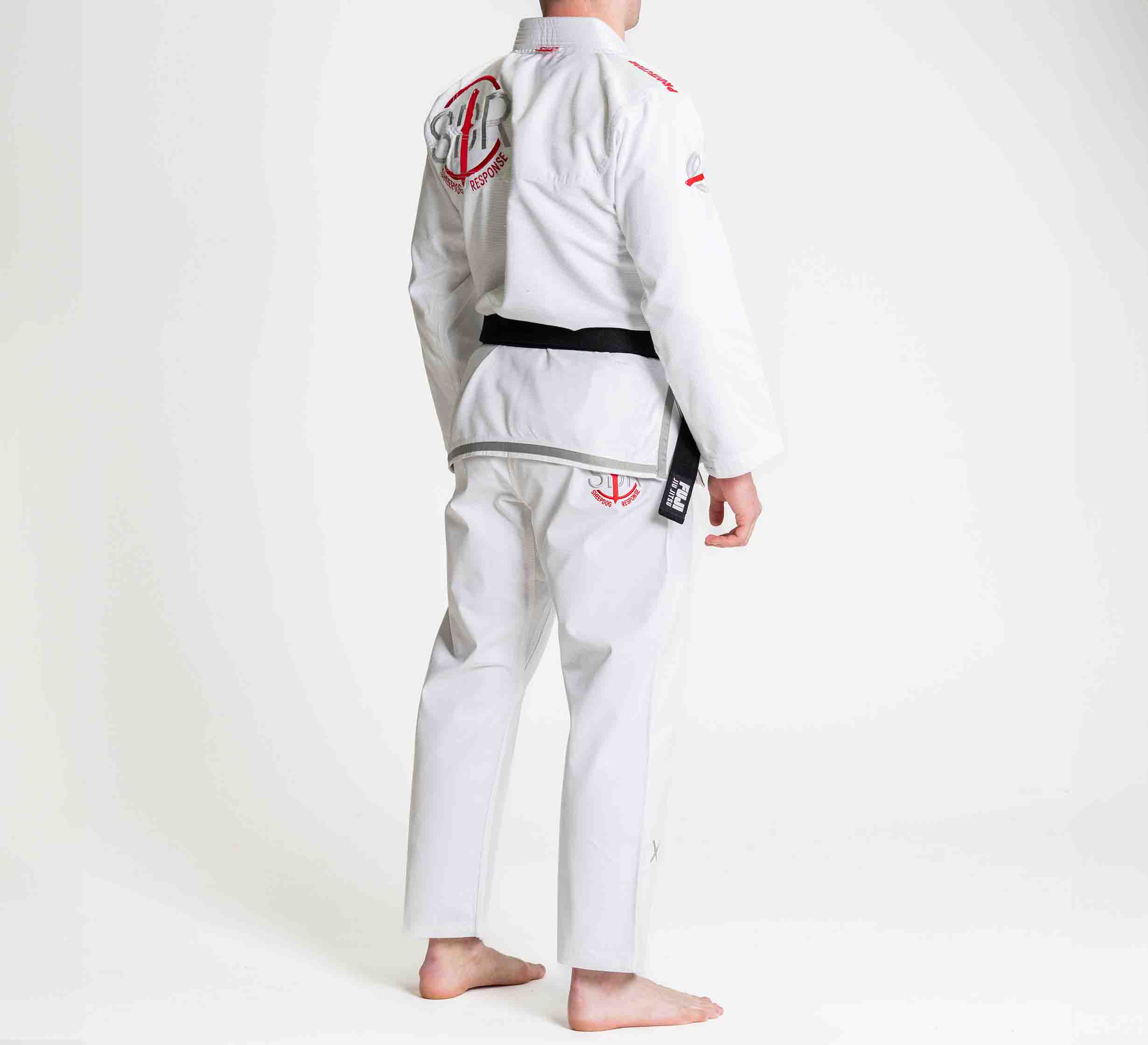 A person stands barefoot on a white background, facing away, wearing the Suparaito BJJ Gi Sheepdog Response Edition White with red patches and a black belt.