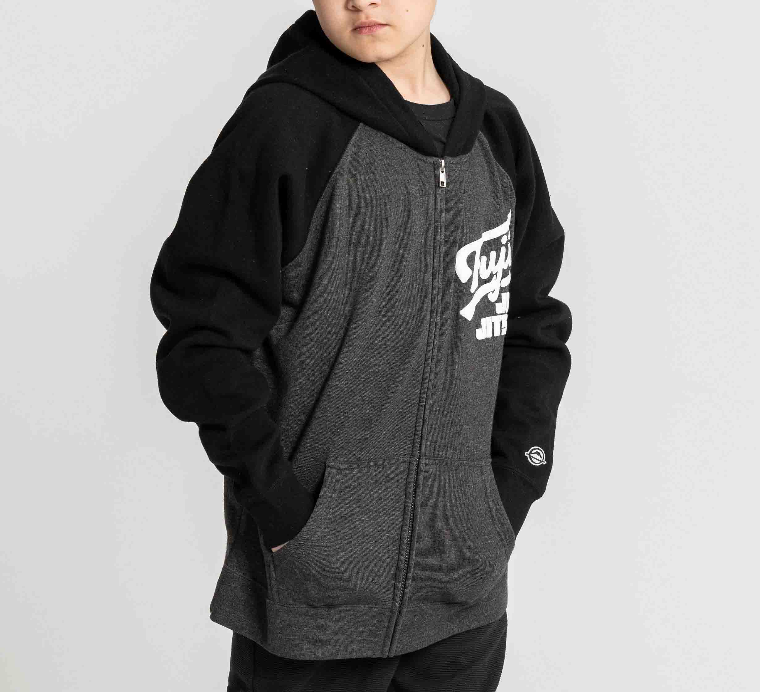 A child wears the Kids Jiu Jitsu Varsity Zip Hoodie in charcoal, featuring front pockets, a zip-up closure, and white text on the chest, standing with hands in pockets against a plain light background.