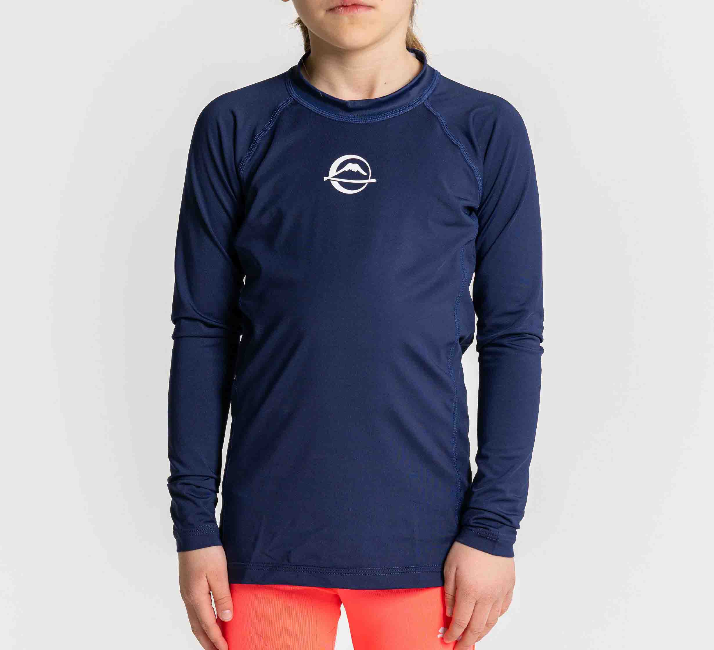 A child wears the Baseline Kids Rashguard Navy with a white chest logo and bright coral shorts, standing against a plain light background.