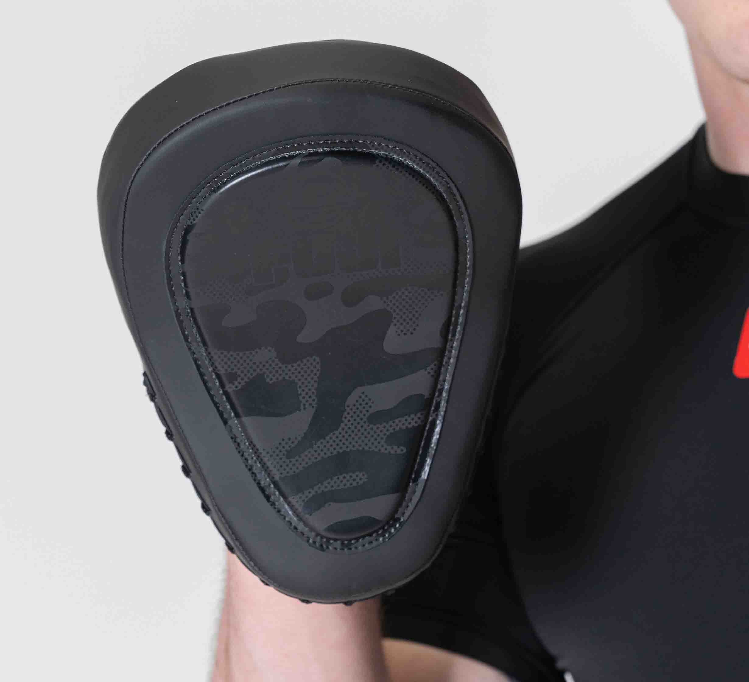 A person in a black shirt holds Raijin Focus Mitts, featuring a black and camouflage design on the front, at chest height against a plain light background.