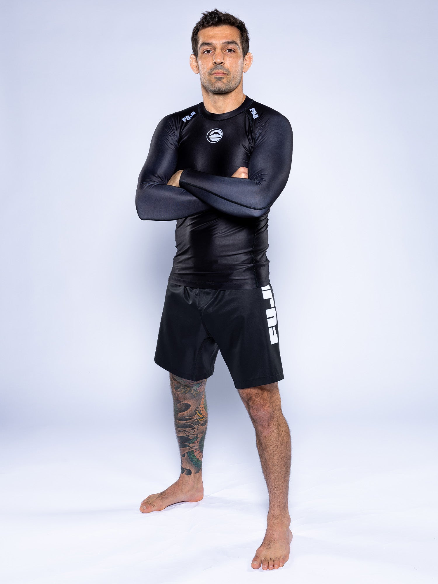 A man with short dark hair and a left leg tattoo stands barefoot with arms crossed, wearing the Baseline 2.0 Long Sleeve Rashguard Black and black FUJI shorts against a plain light background.