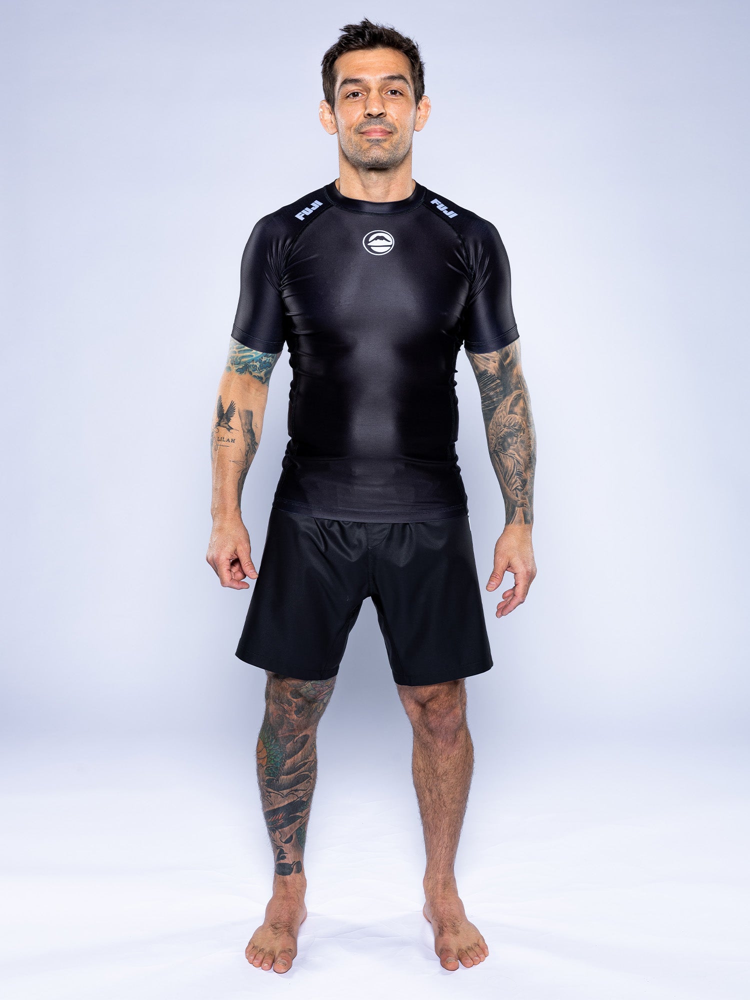 A tattooed man stands barefoot on a white background, wearing the Baseline 2.0 Short Sleeve Rashguard in black and matching athletic shorts, looking at the camera with a neutral expression.