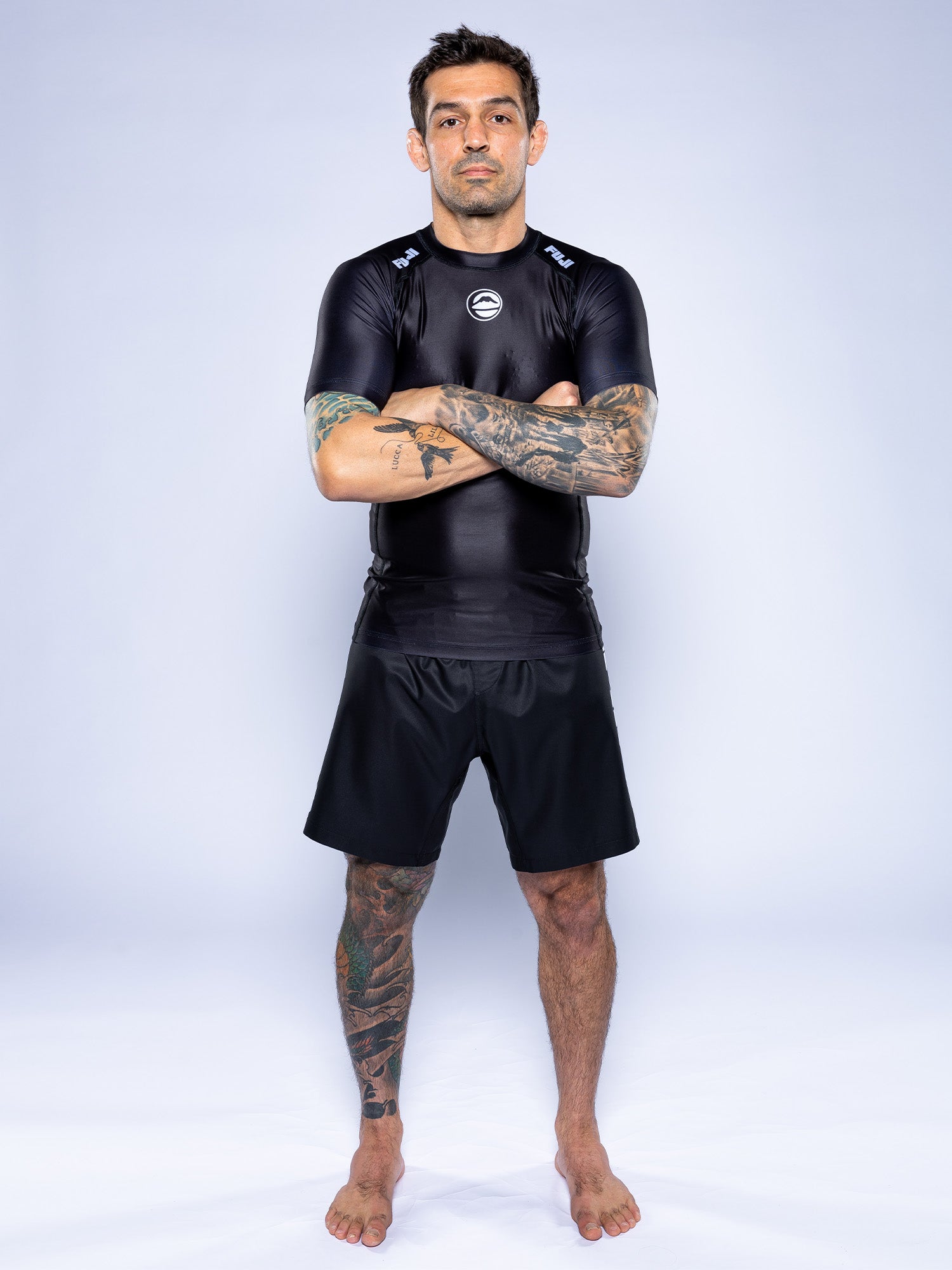 A tattooed man stands barefoot on a light background, wearing the Baseline 2.0 Short Sleeve Rashguard Black and black shorts, arms crossed.