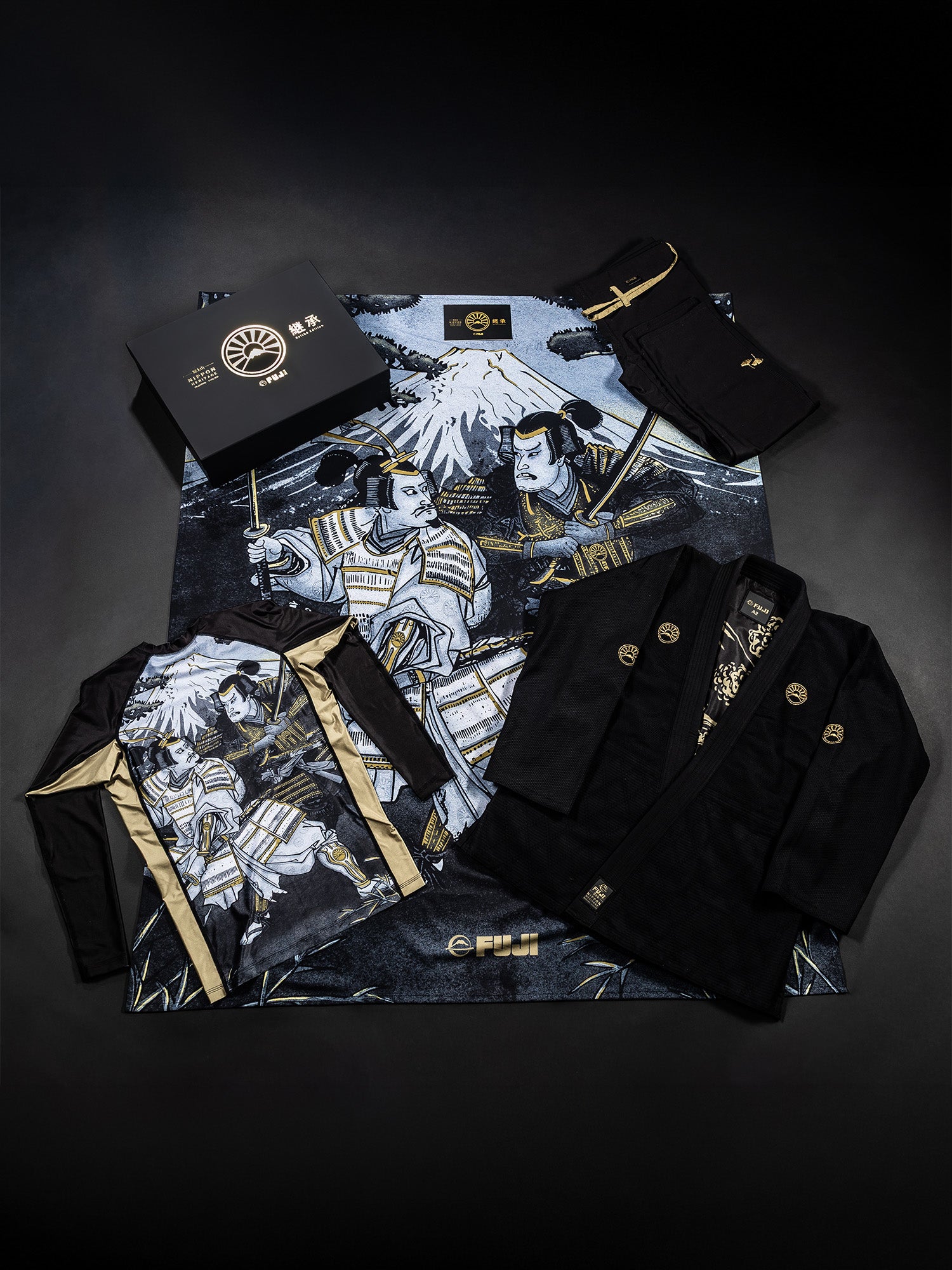 Flat lay of the Heritage Keishō Edition Bundle, featuring a black martial arts gi, matching pants, a folded belt, a presentation box, and an intricately detailed mat adorned with samurai warriors and Mount Fuji on a dark background.