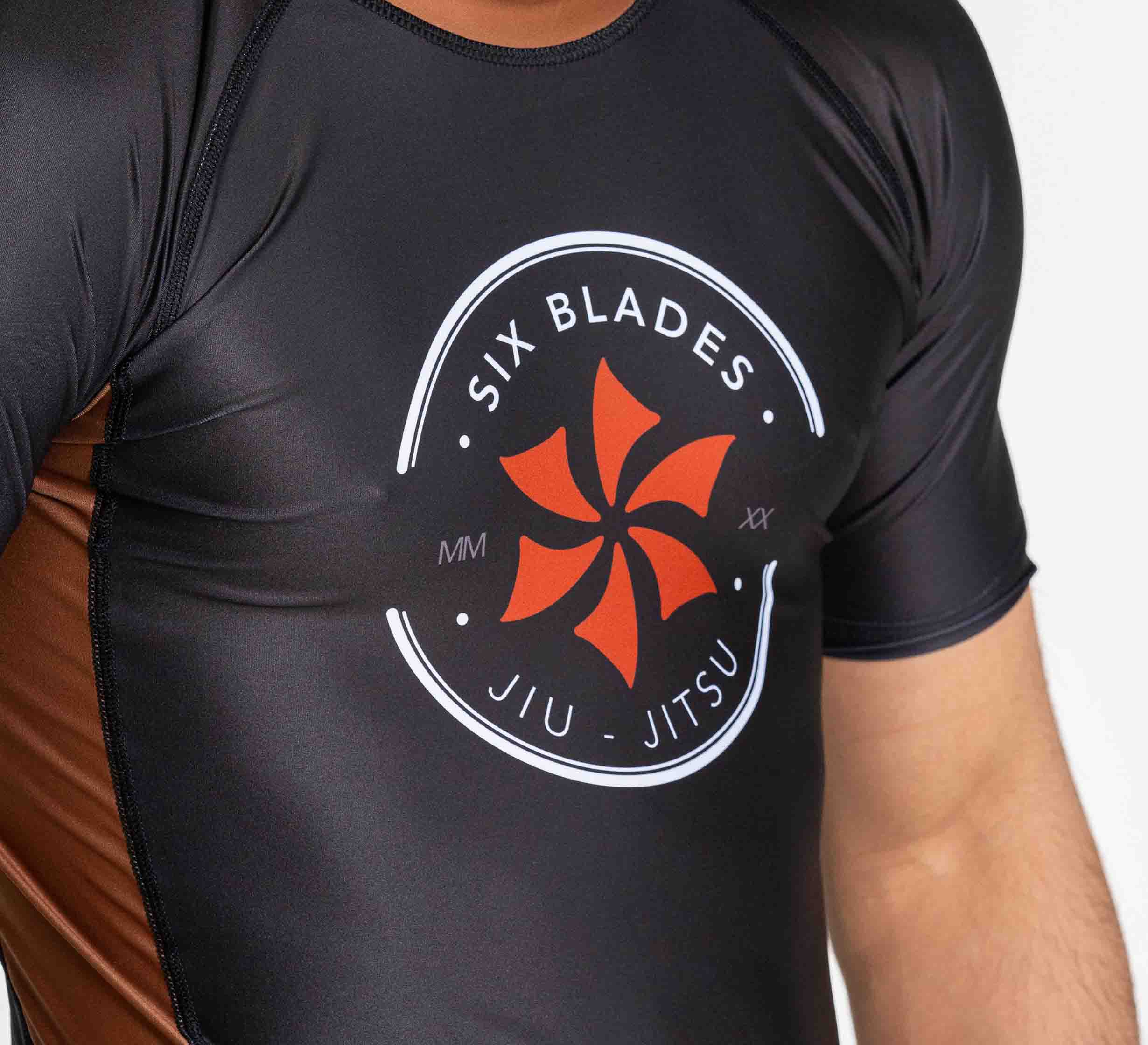 Someone wearing the Six Blades Short Sleeve Rashguard Brown, which features a black and brown athletic design with the Six Blades Jiu Jitsu logo—a red six-bladed emblem and white text—on the chest.