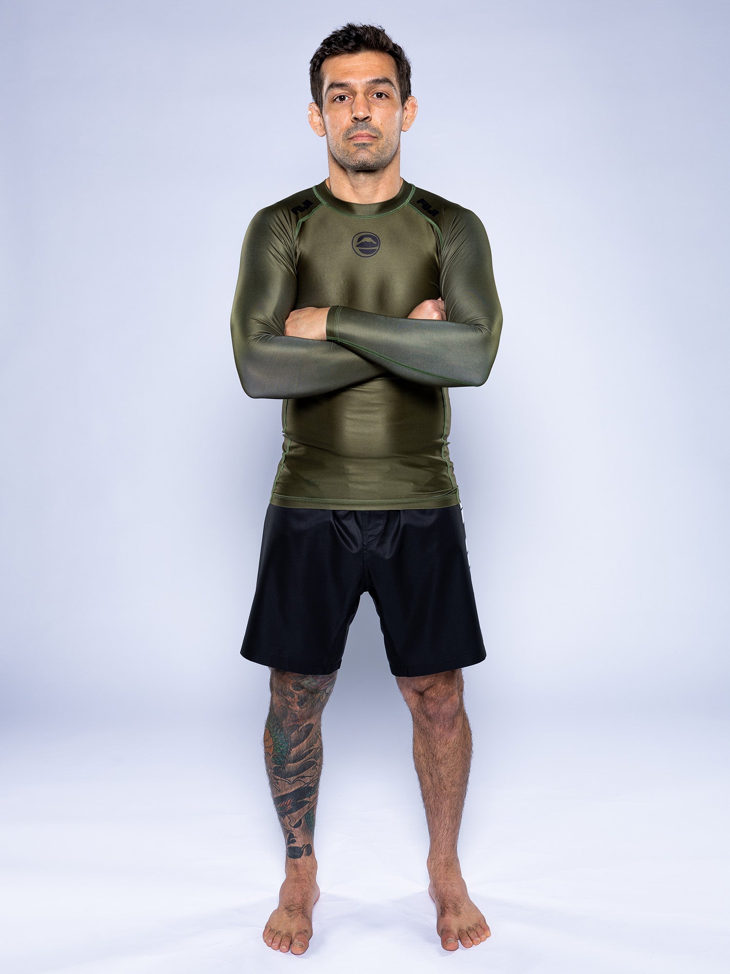 A man stands barefoot on a white background with arms crossed, wearing the Baseline 2.0 Long Sleeve Rashguard in Military Green and black shorts. A tattoo is visible on his left leg.