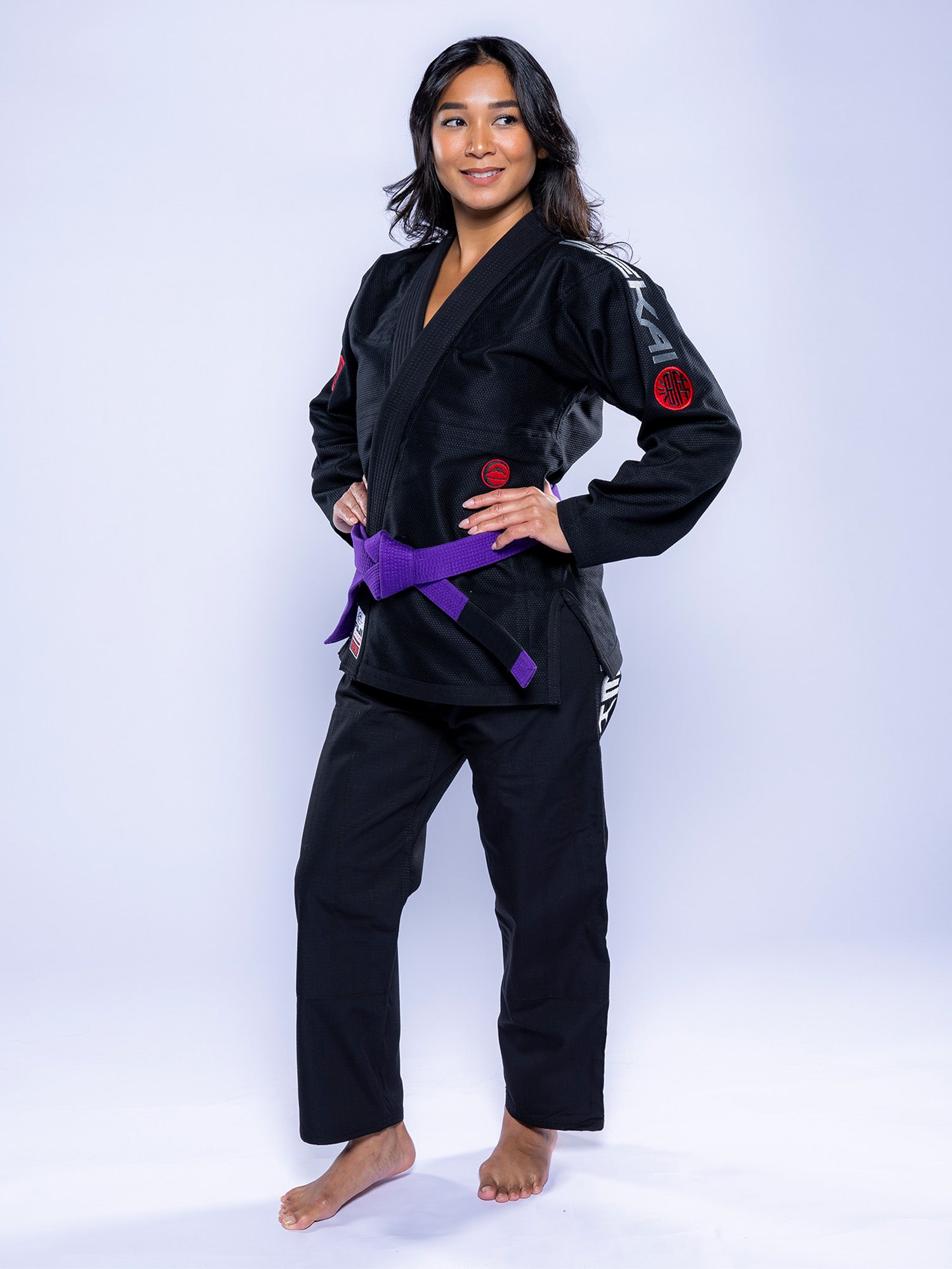 A woman stands barefoot on a white background, wearing the Womens SEKAI 3.0 BJJ Gi Black Grey and a purple belt. She faces forward confidently with hands on hips and a relaxed expression.