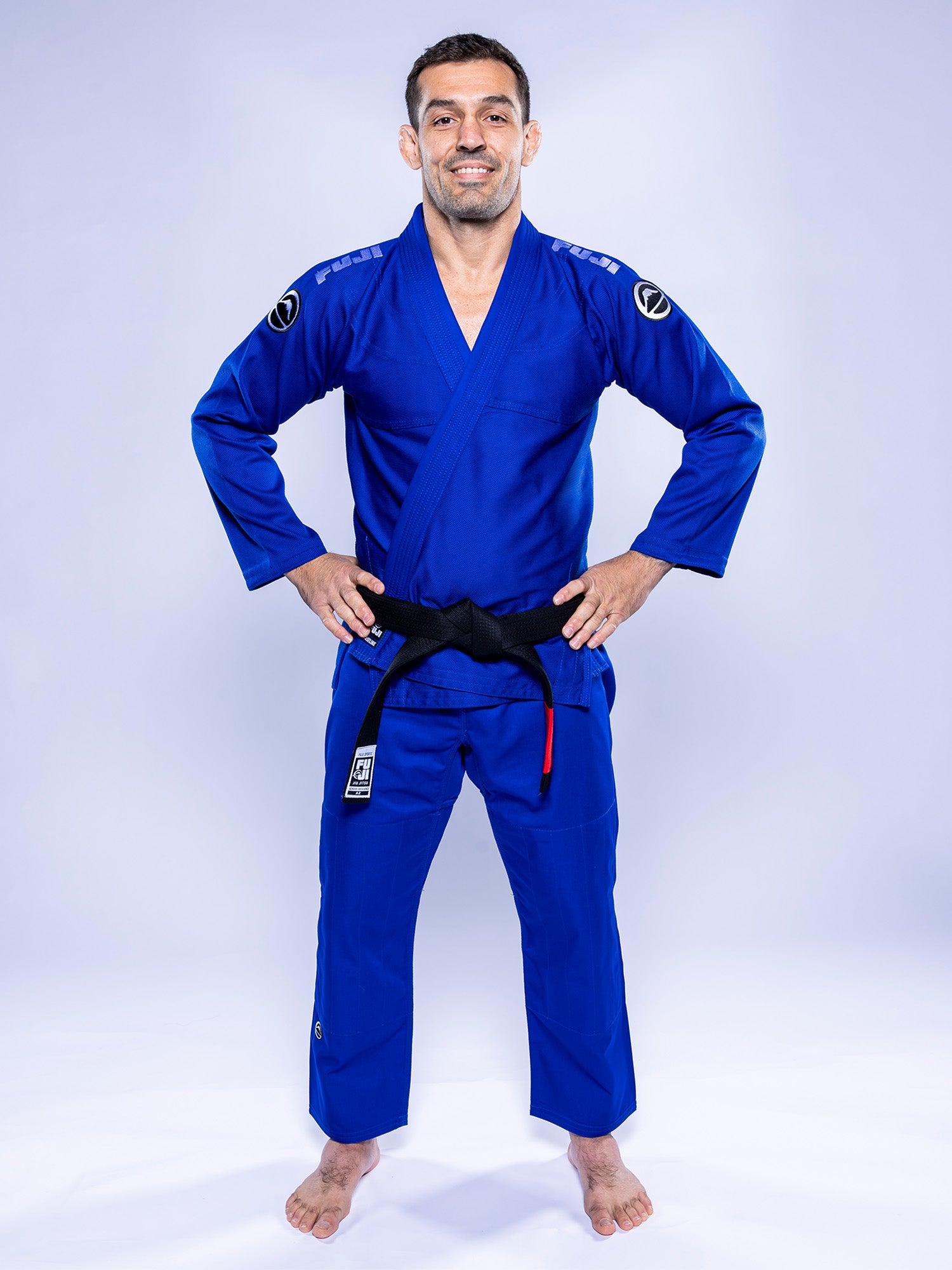 A man stands barefoot on a white background, smiling and wearing the Baseline 2.0 BJJ Gi Blue White with a black belt. His hands are on his hips, and he looks directly at the camera.
