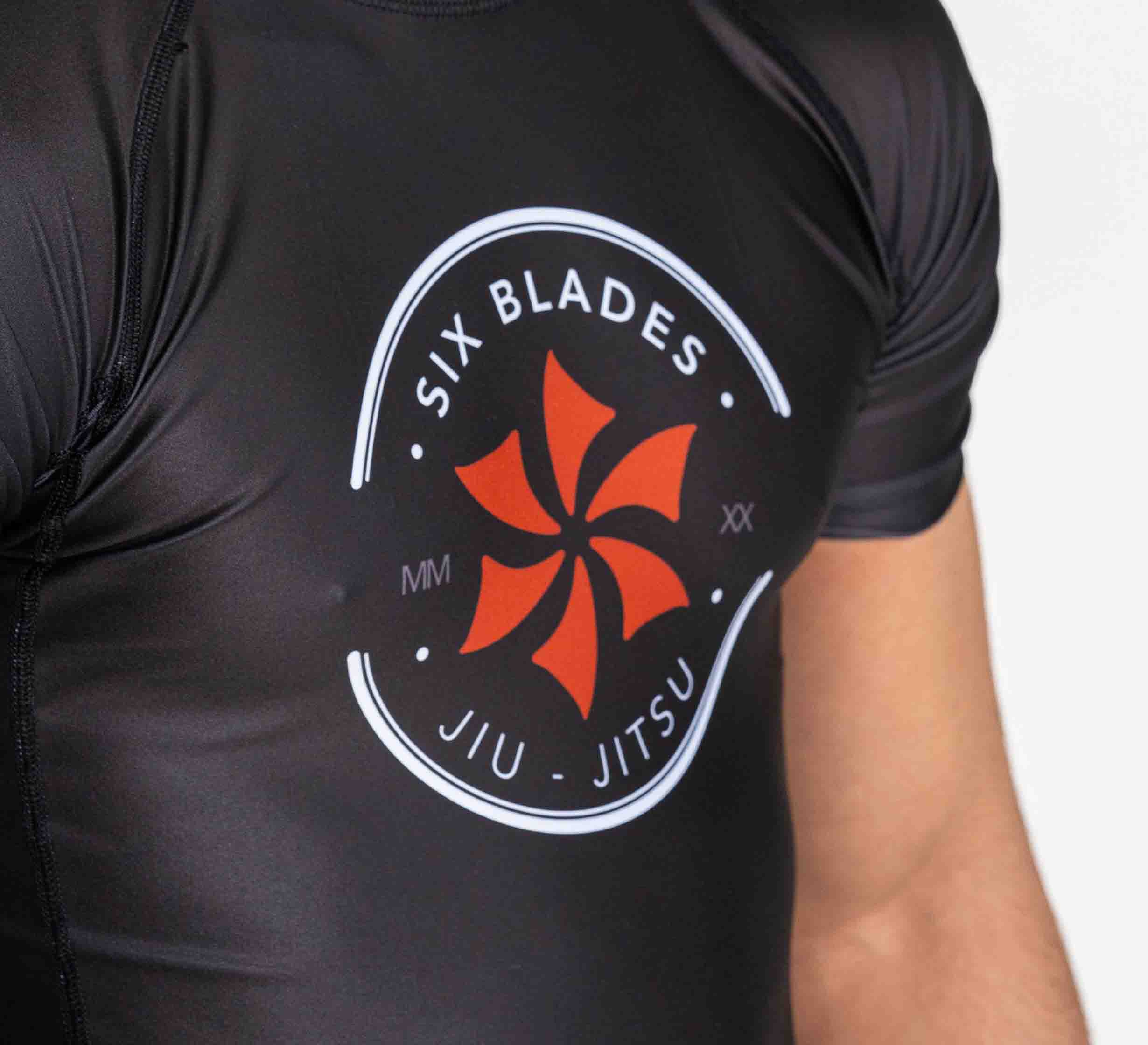A person wears the Six Blades Short Sleeve Rashguard Black featuring a circular Six Blades Jiu Jitsu logo and a red six-bladed pinwheel design on the chest.