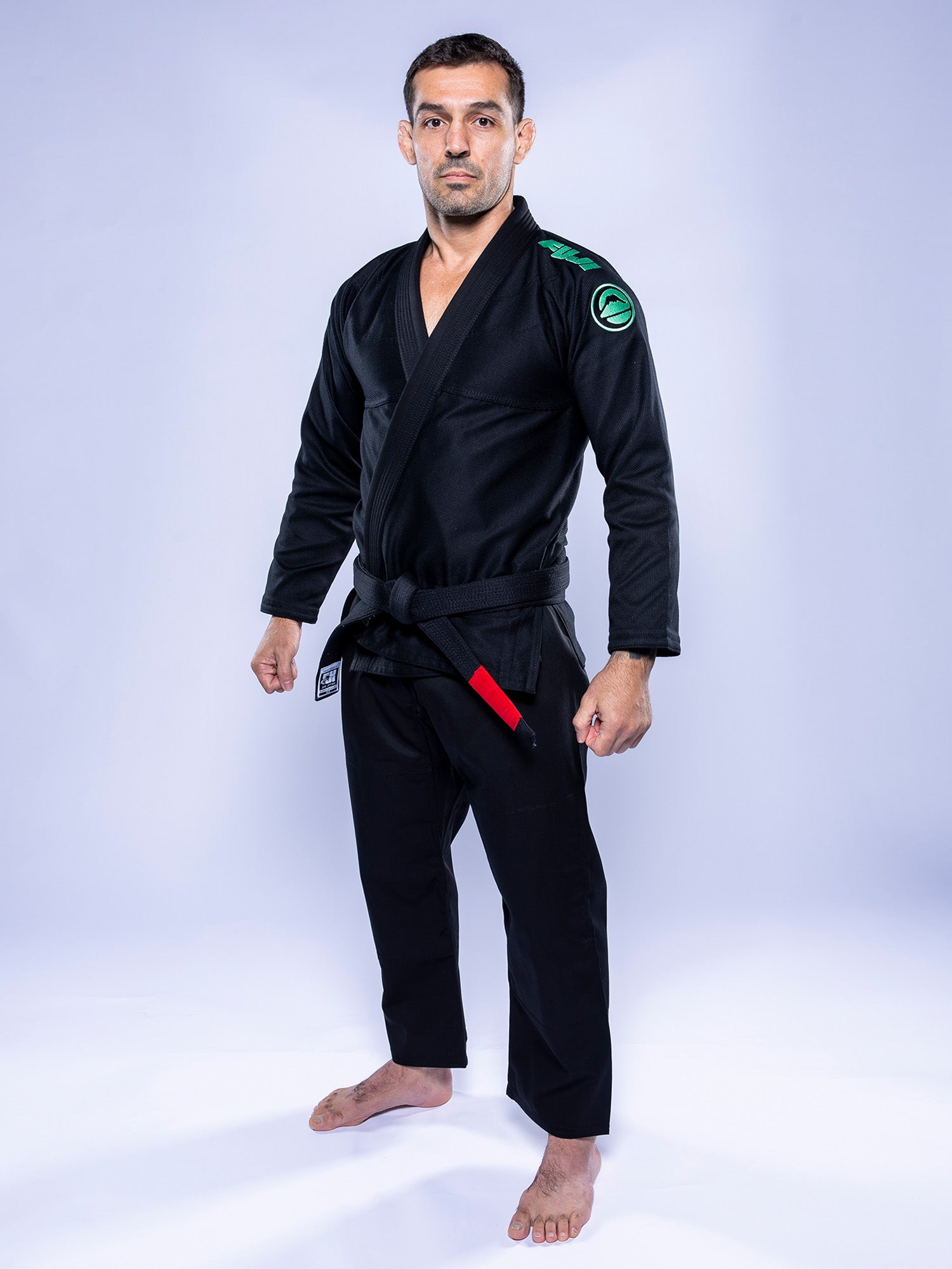 A man faces forward on a white floor, wearing the Baseline 2.0 BJJ Gi Black Mint with a black belt. The gi features a mint green patch on the shoulder. He stands barefoot with a neutral expression against a plain light background.