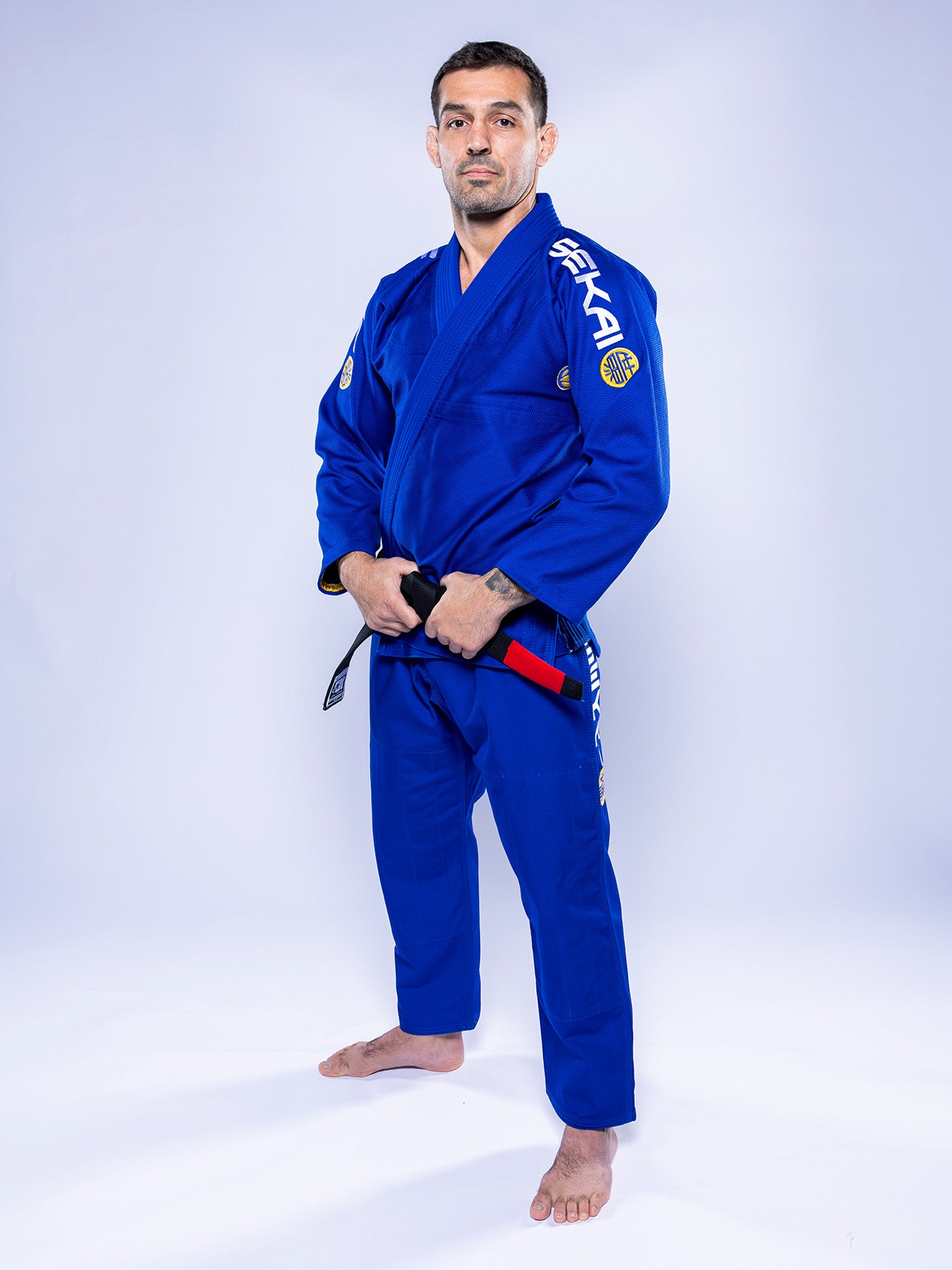 A man wearing the SEKAI 3.0 BJJ Gi Blue Yellow and a black belt stands barefoot on a white background, confidently looking at the camera with his hands gripping his belt.