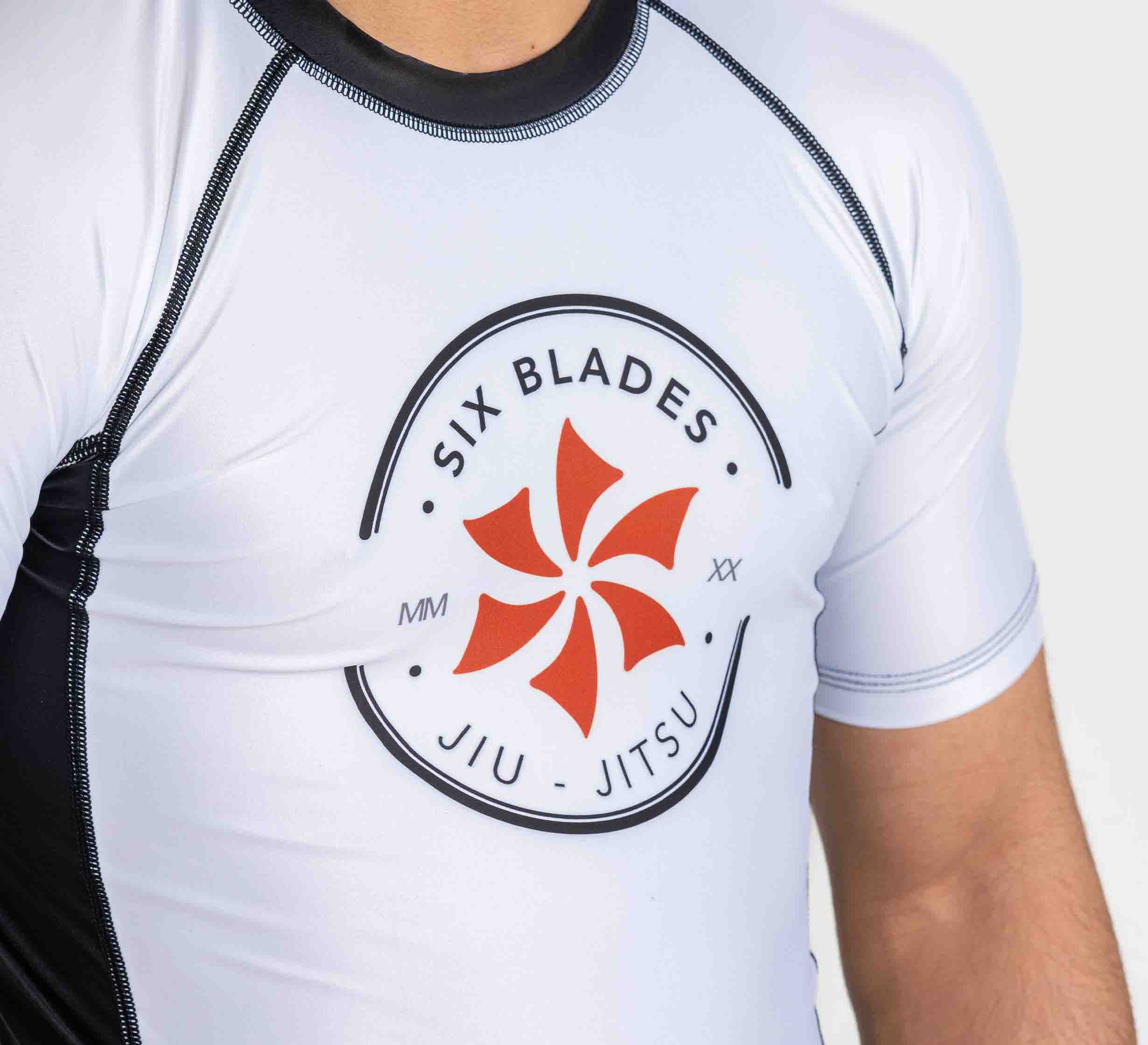 A child wears the Kids Six Blades Short Sleeve Rashguard White, a white athletic top with black trim, red pinwheel logo, and "Six Blades Jiu Jitsu" text on the chest.
